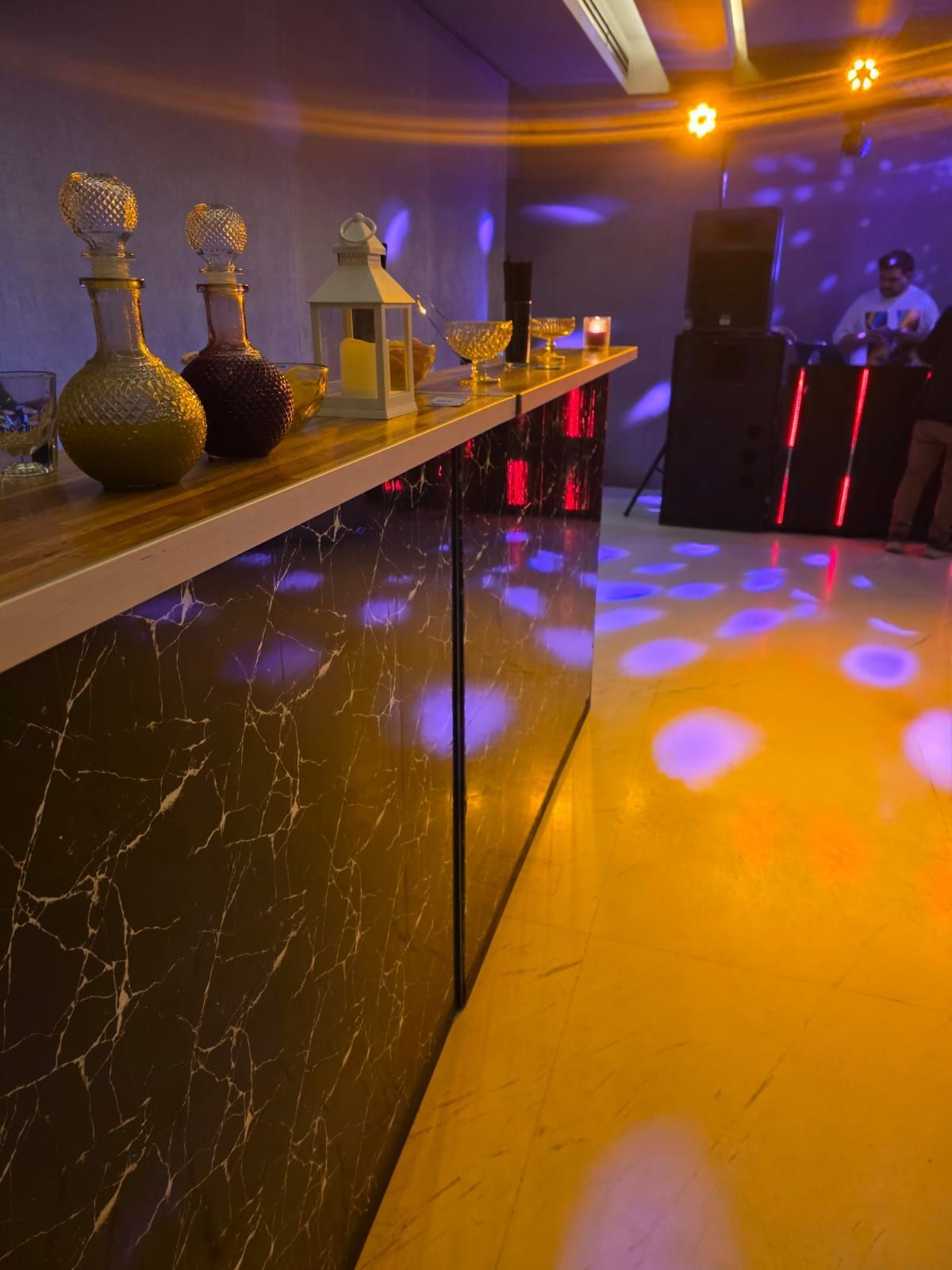 Salón con iluminación tenue, jarrones decorativos en una estantería y focos azul-violeta en el suelo y las paredes.