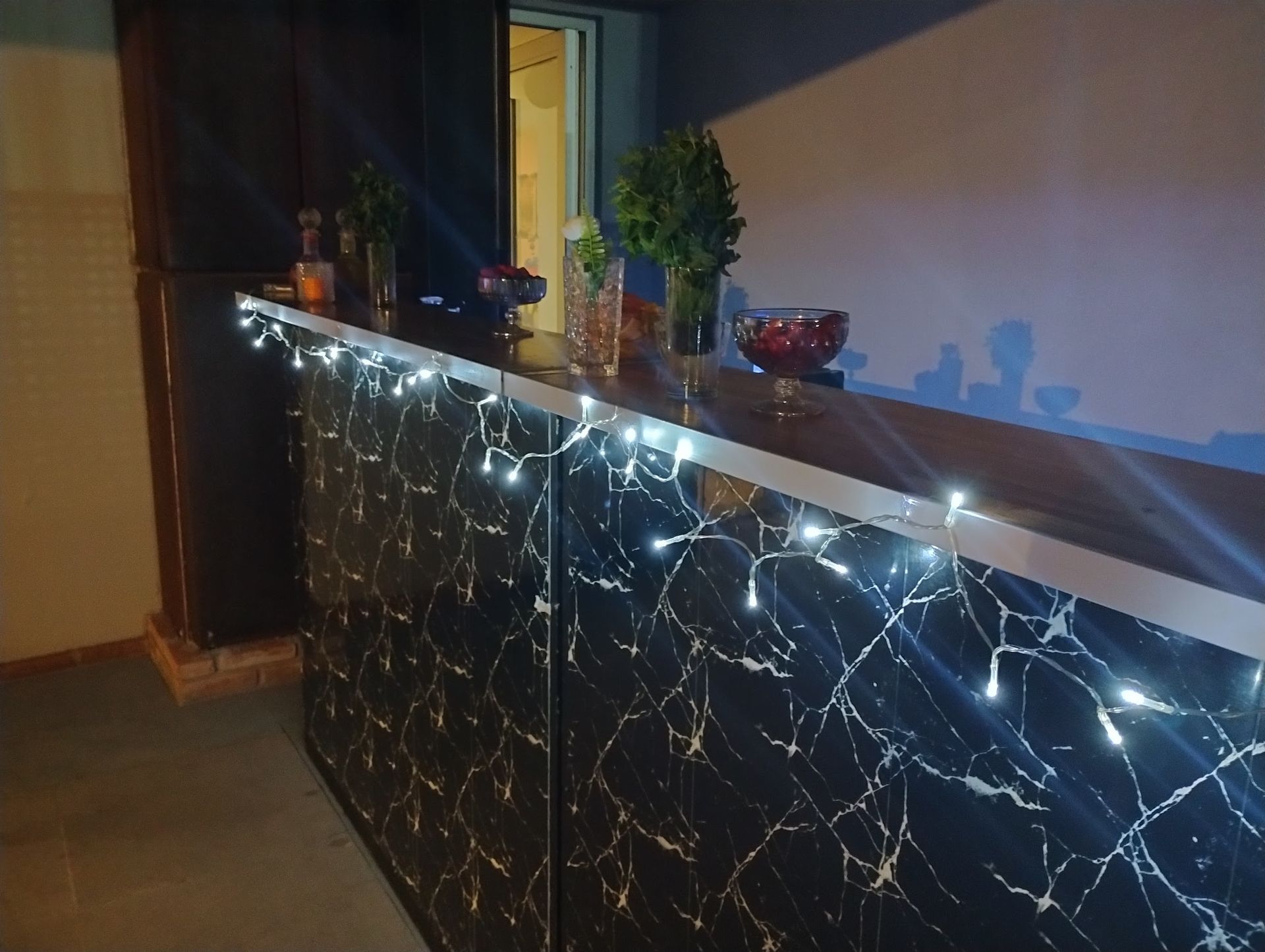 Un mostrador de mármol oscuro con guirnaldas de luces blancas y pequeñas plantas en un interior con poca luz.