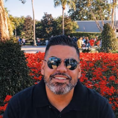 Man smiling in sunglasses, seated outdoors with red flowers and trees in the background