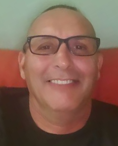 Smiling person wearing black glasses, seated against a red couch and pale green wall.