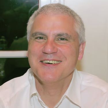 Smiling man in a white shirt, seated indoors against a softly lit green background