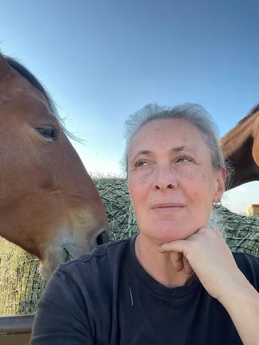Woman outdoors with a brown horse nuzzling her cheek under a clear blue sky