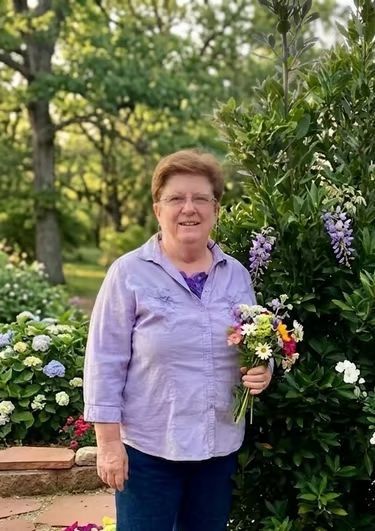 Person standing outdoors in a garden, holding a bouquet of flowers, wearing a lavender shirt.