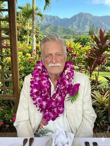 Man wearing a white shirt and purple lei seated at a table with tropical mountains in the background