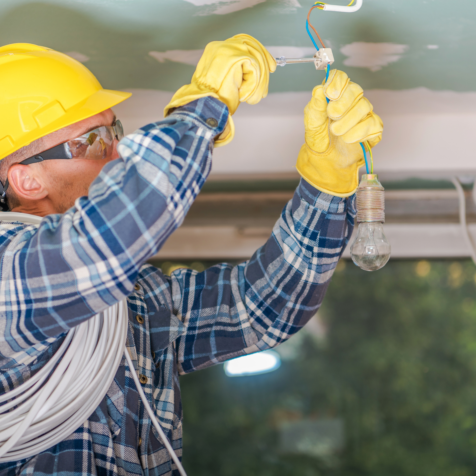 Electrician in a hard hat and gloves connecting wires to a light fixture in a room.