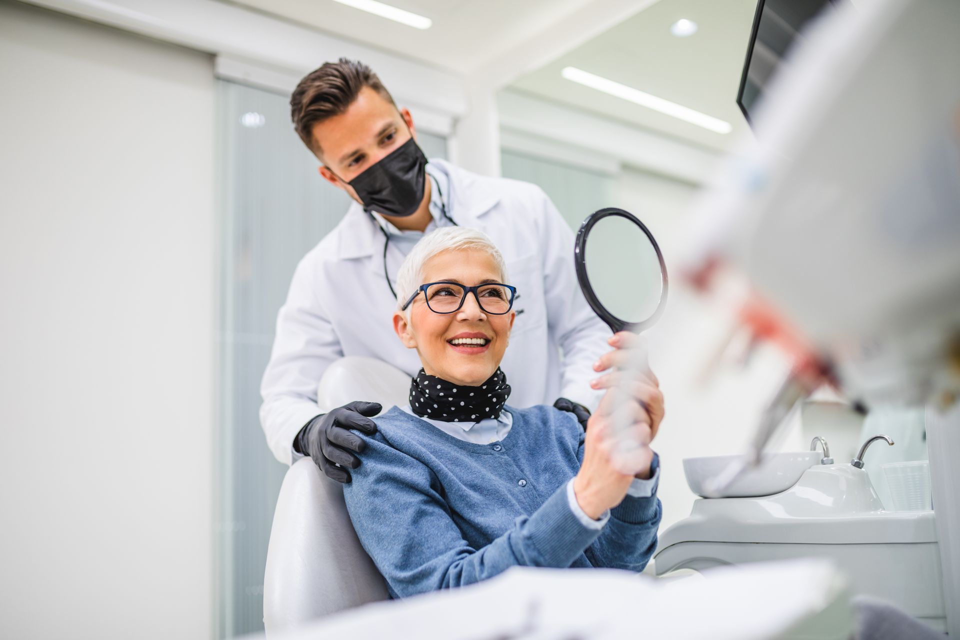 Dentist in white coat and mask smiles with seated patient holding a mirror in a bright dental clinic Dentist in white coat and mask smiles with seated patient holding a mirror in a bright dental clinic