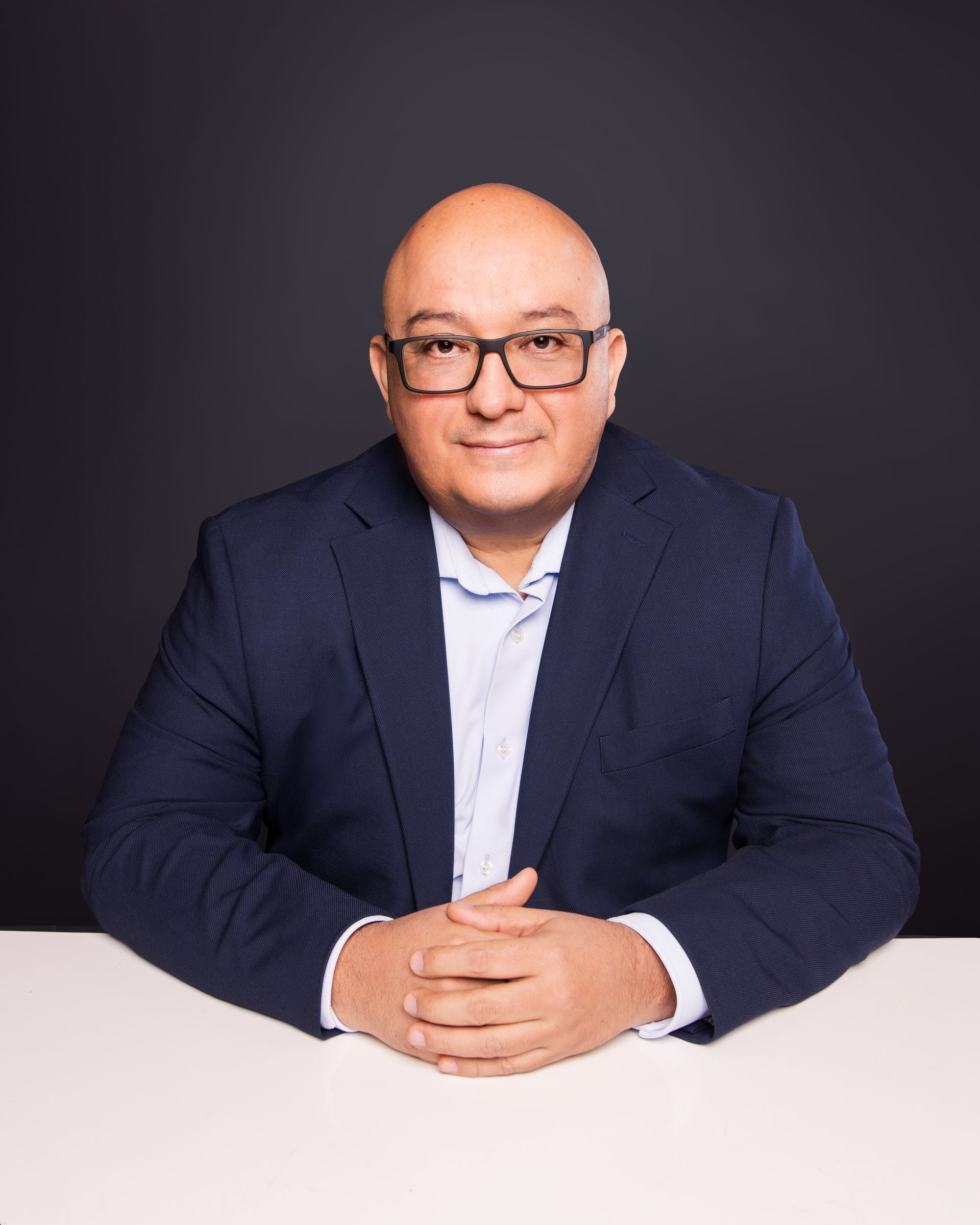 Man seated at a table, wearing a navy blazer and glasses, against a dark studio background.