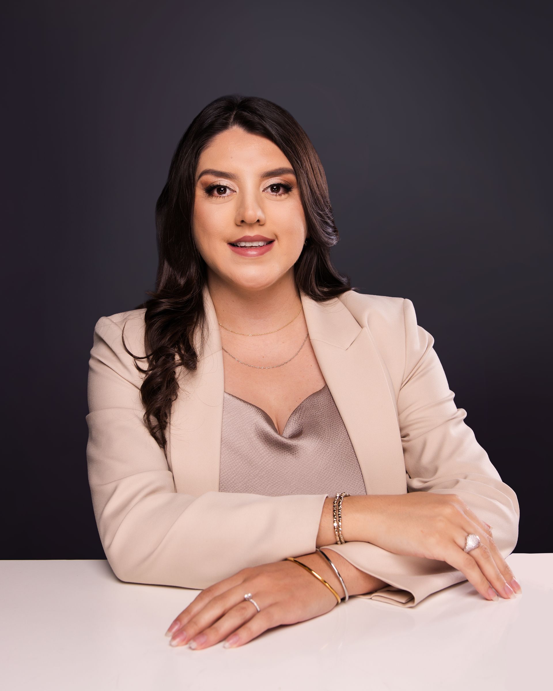 Woman seated at a table against a dark studio backdrop, wearing a beige blazer and smiling at the camera