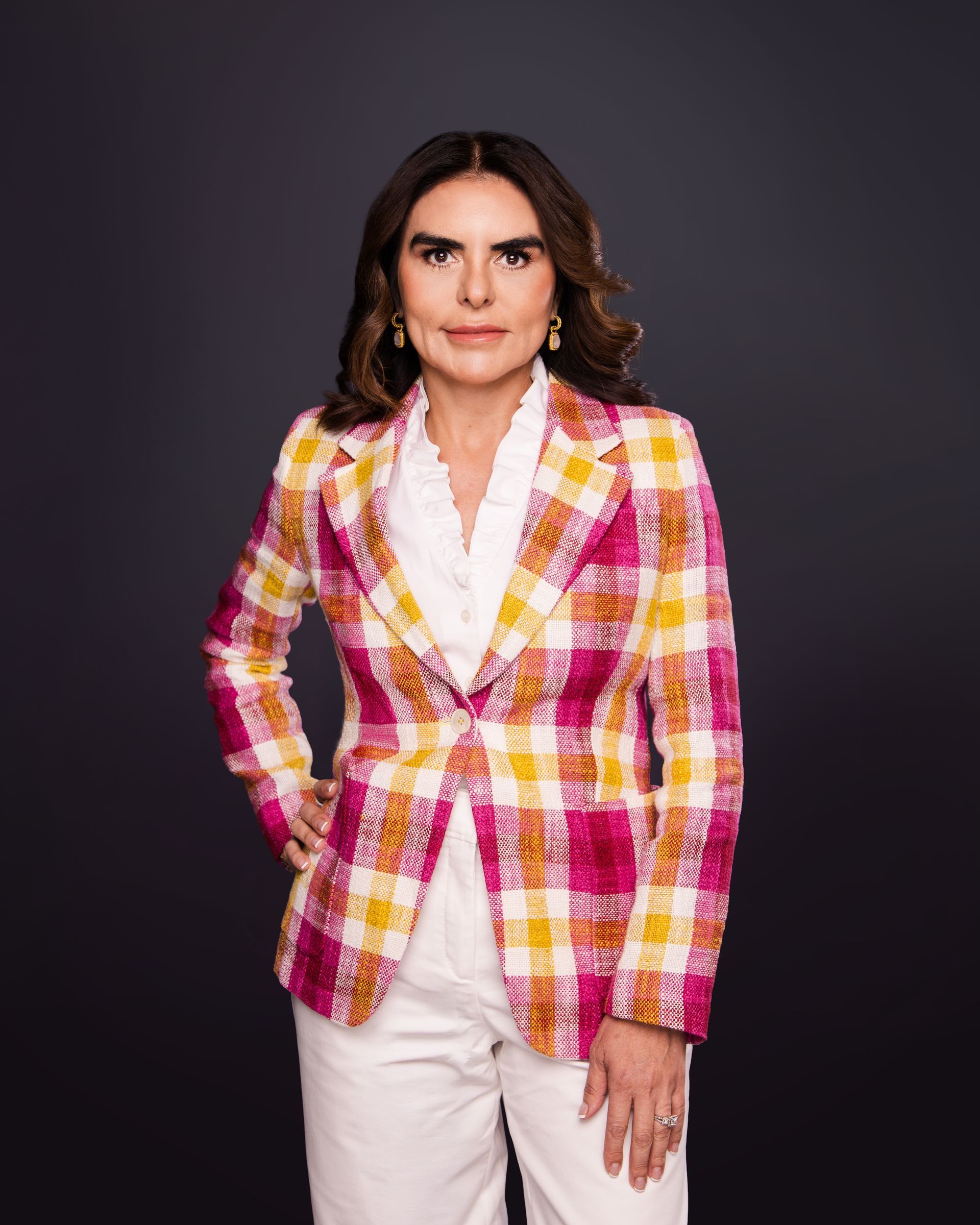 Woman in a pink-and-yellow plaid blazer and white pants standing against a dark gray backdrop.