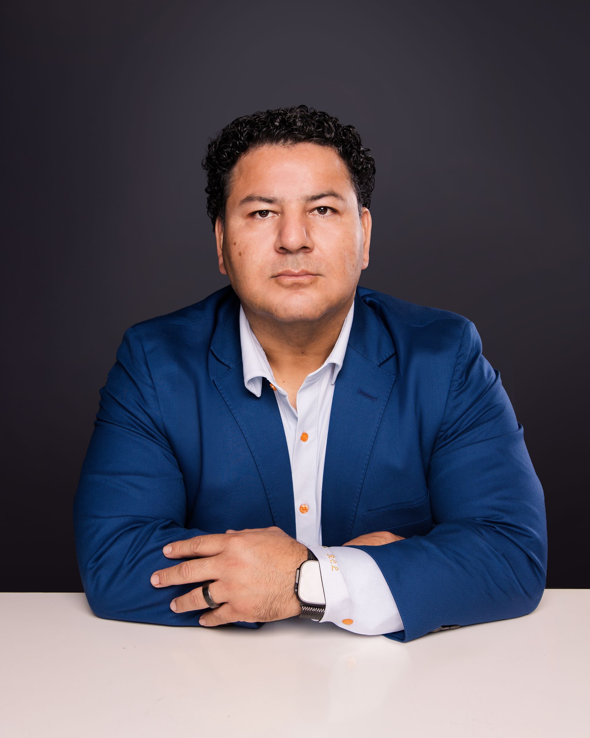 Portrait of a man in a blue blazer seated at a table against a dark background.