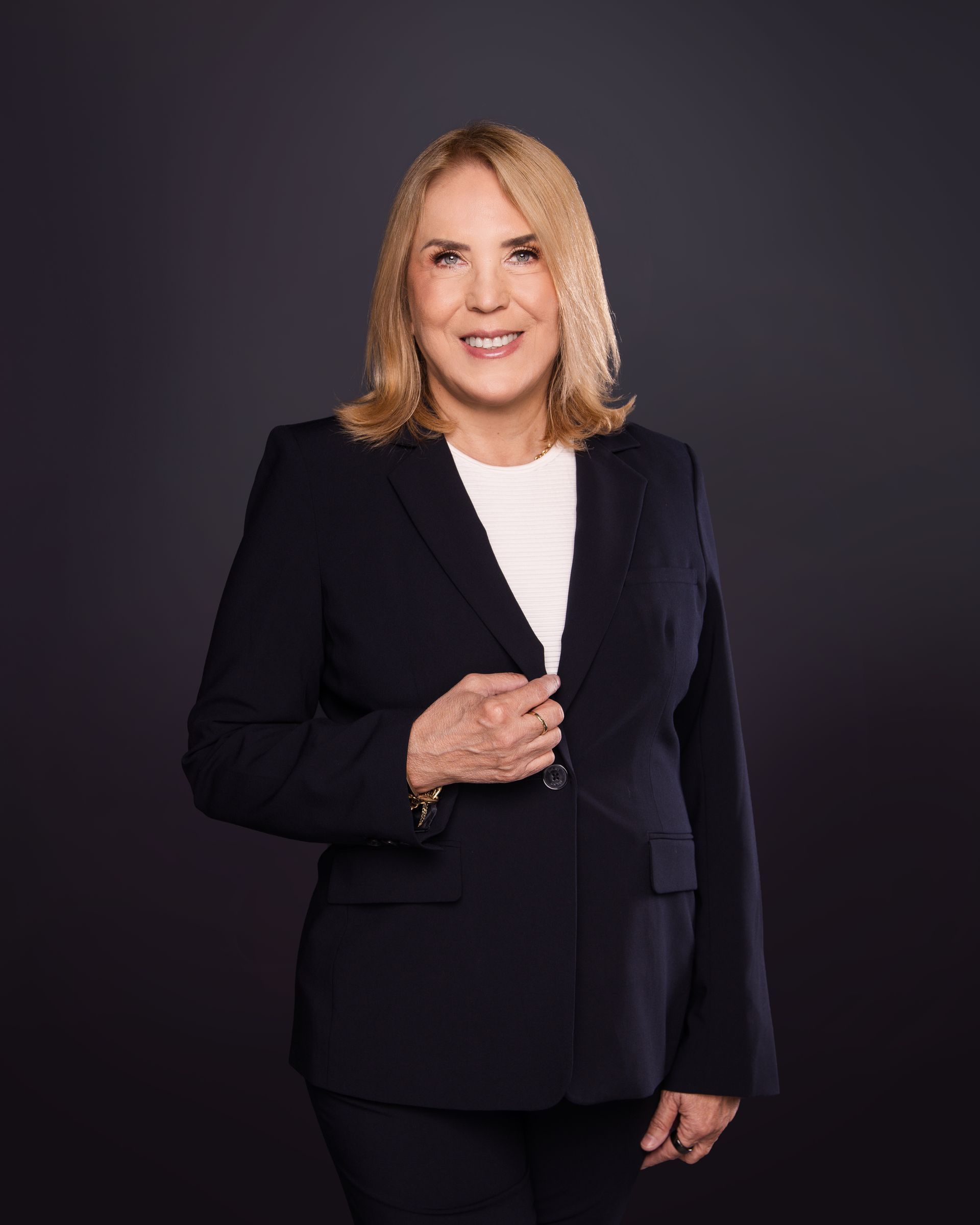 Professional portrait of a woman in a black blazer standing against a dark studio background