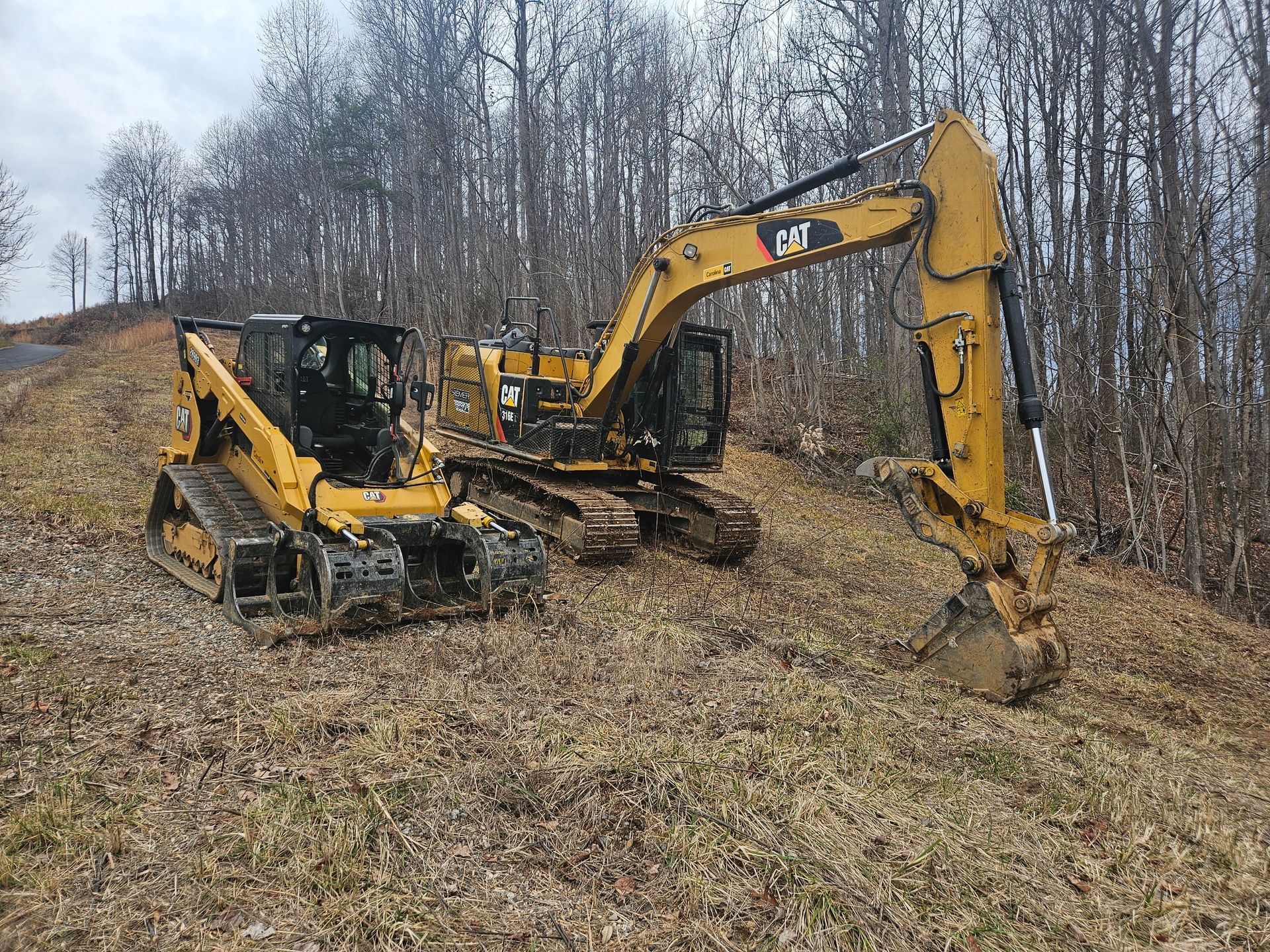 Yellow Caterpillar excavator and skid steer on a grassy hillside near a treeline.
