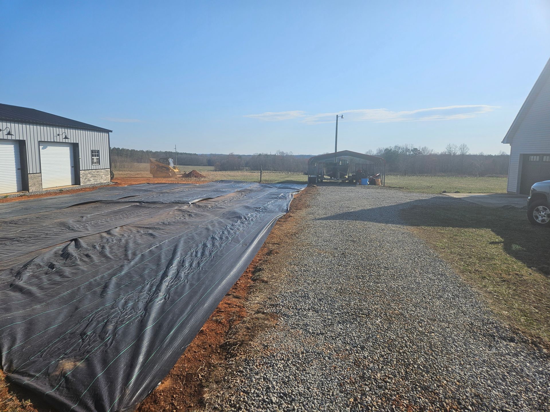 Black tarp laid along a dirt path, gravel driveway to the right, buildings in the background. Bright sky.
