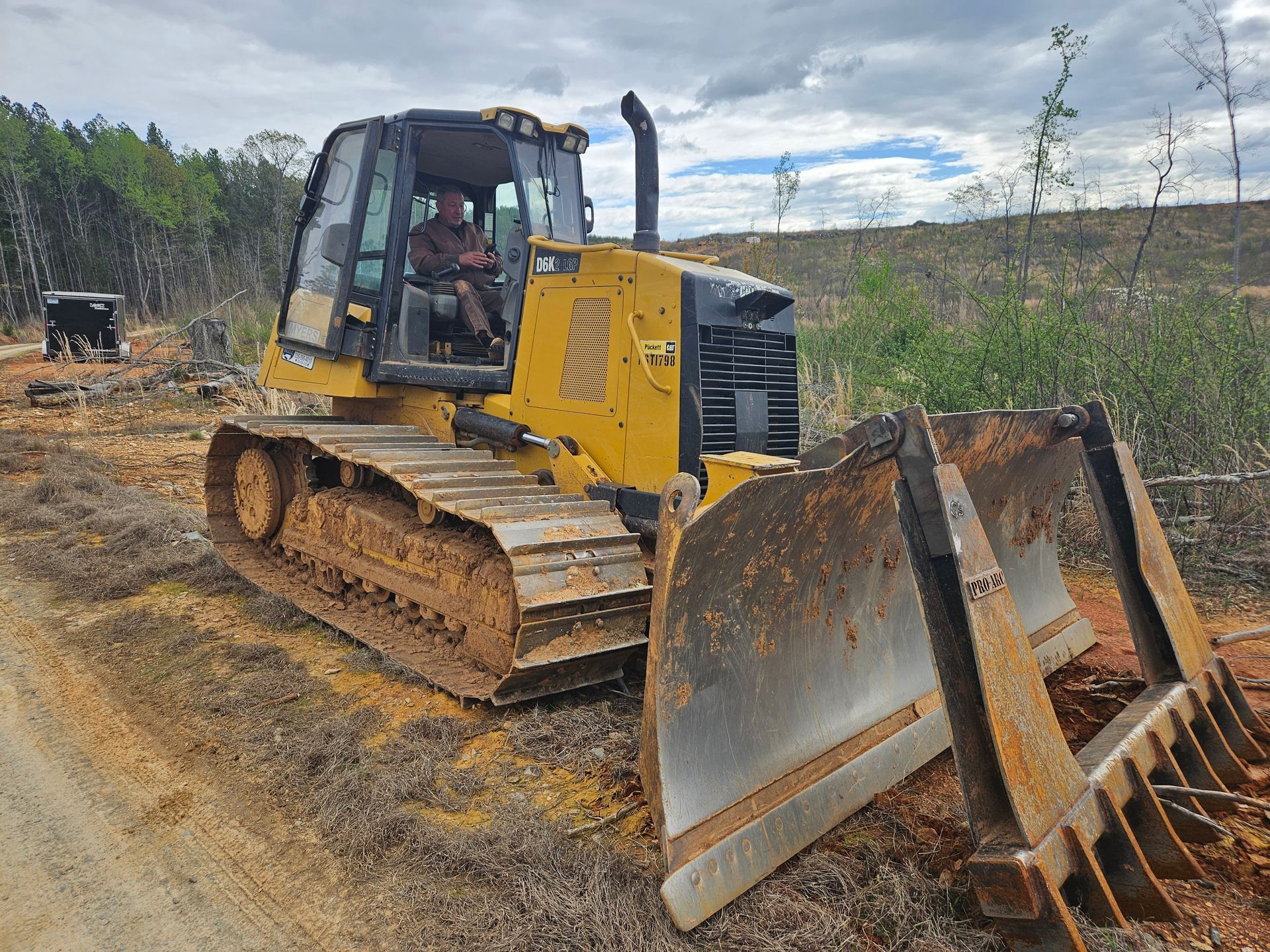 Yellow bulldozer with operator in cab, clearing land with exposed dirt and felled trees.