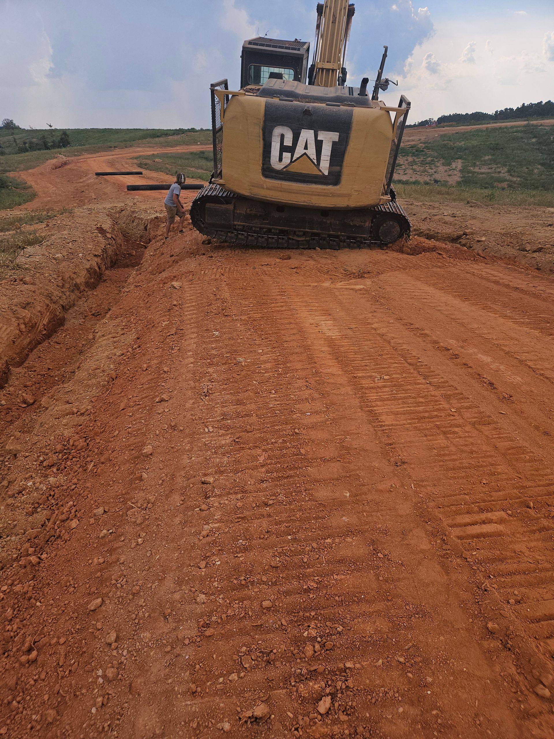 Yellow CAT excavator on a dirt road, grading the soil. A person stands nearby.