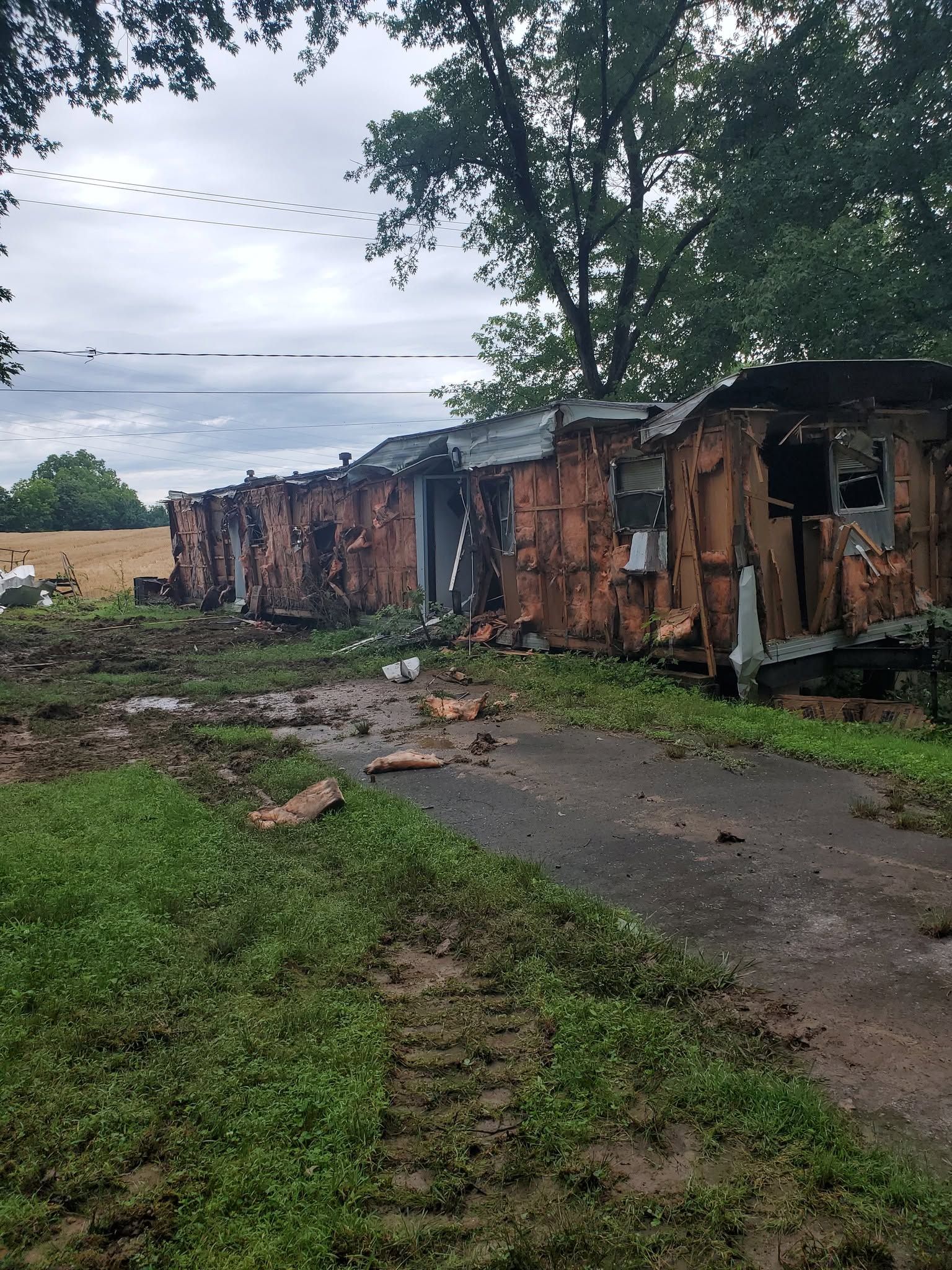 Damaged row of rusted buildings beside a muddy path under cloudy sky, with trees and scattered debris