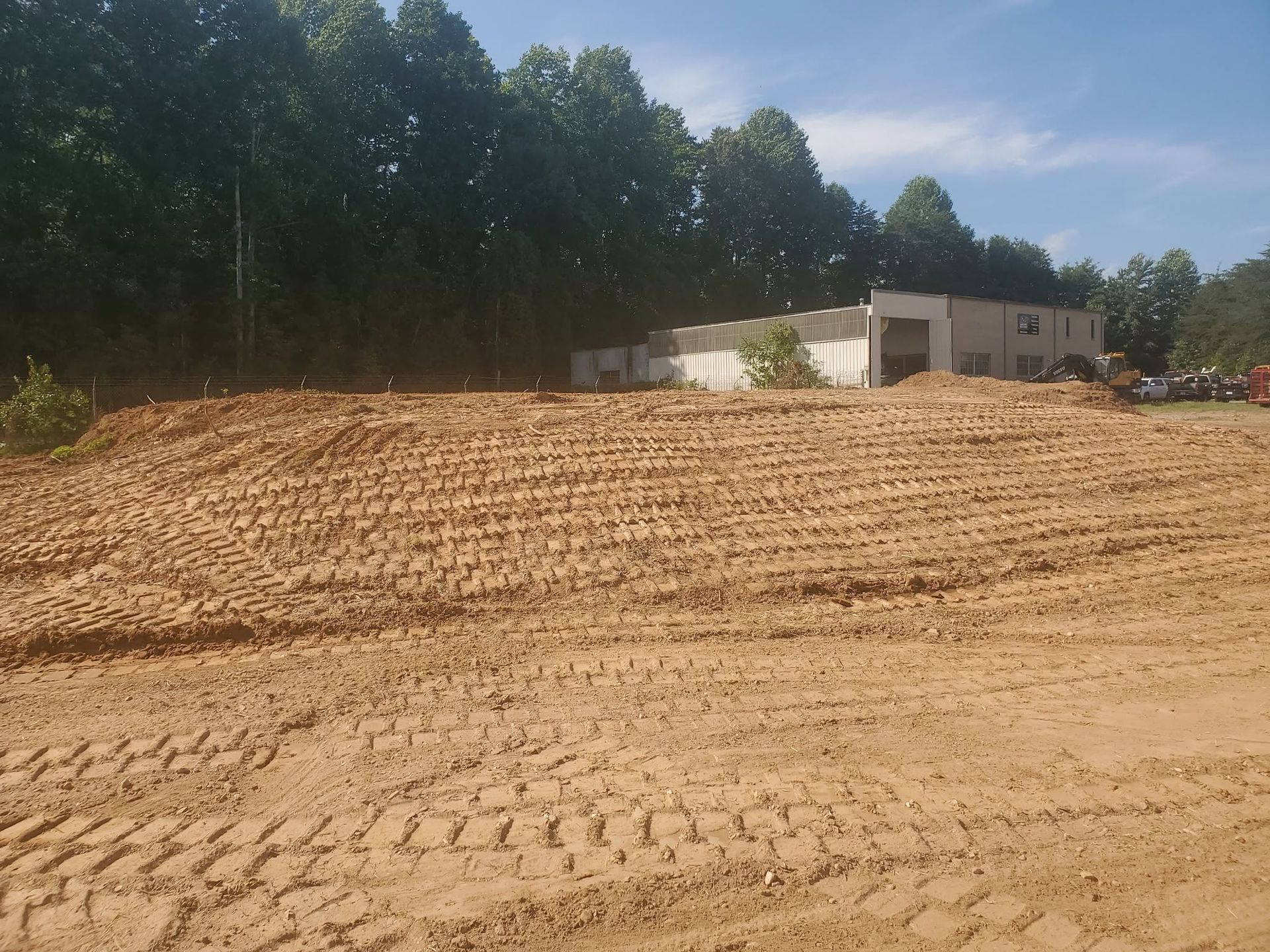 Cleared dirt lot with tire tracks, trees along the left, and a small white building in the background