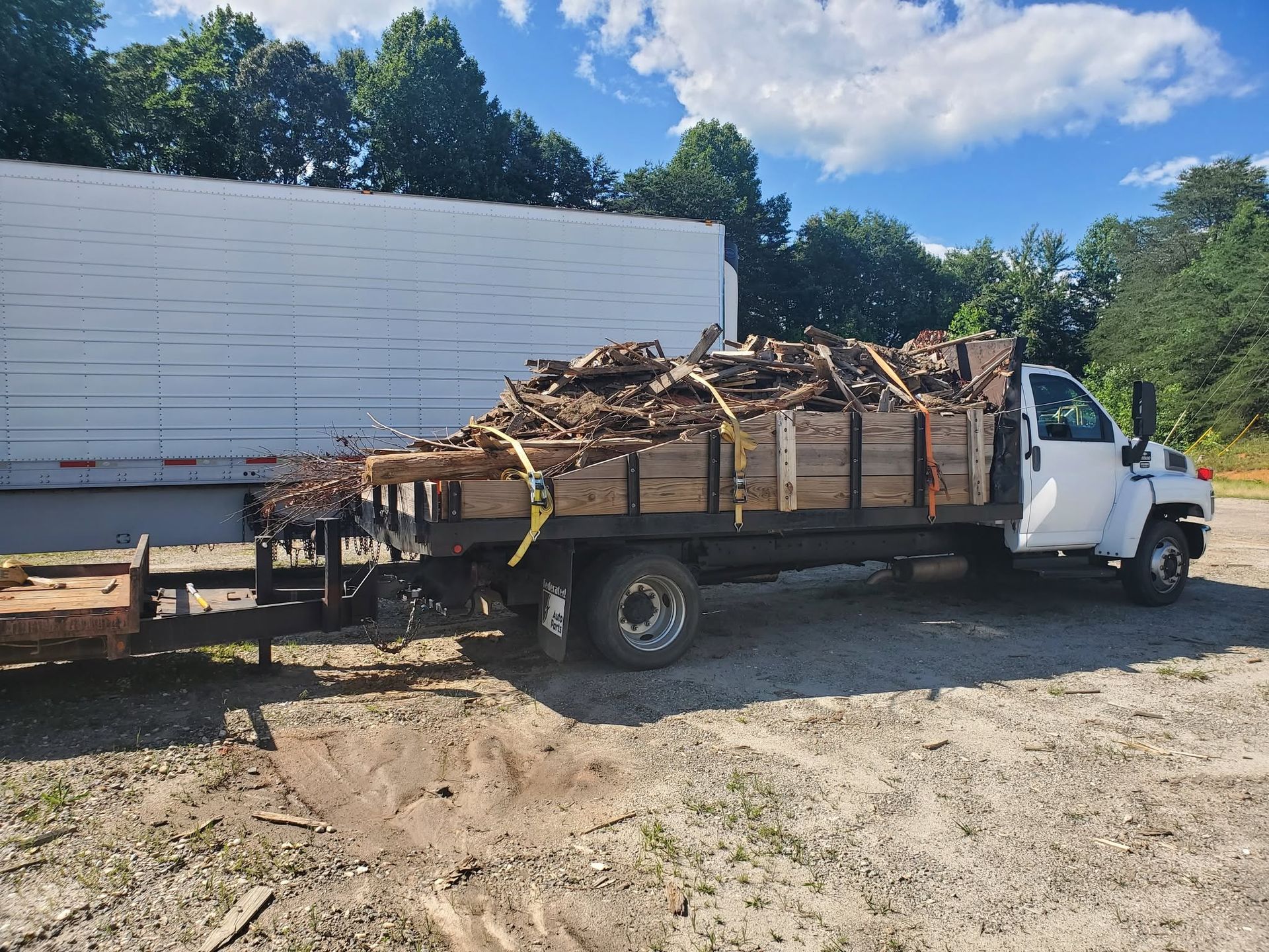 White flatbed truck loaded with debris parked on a dirt lot beside a white building under a blue sky