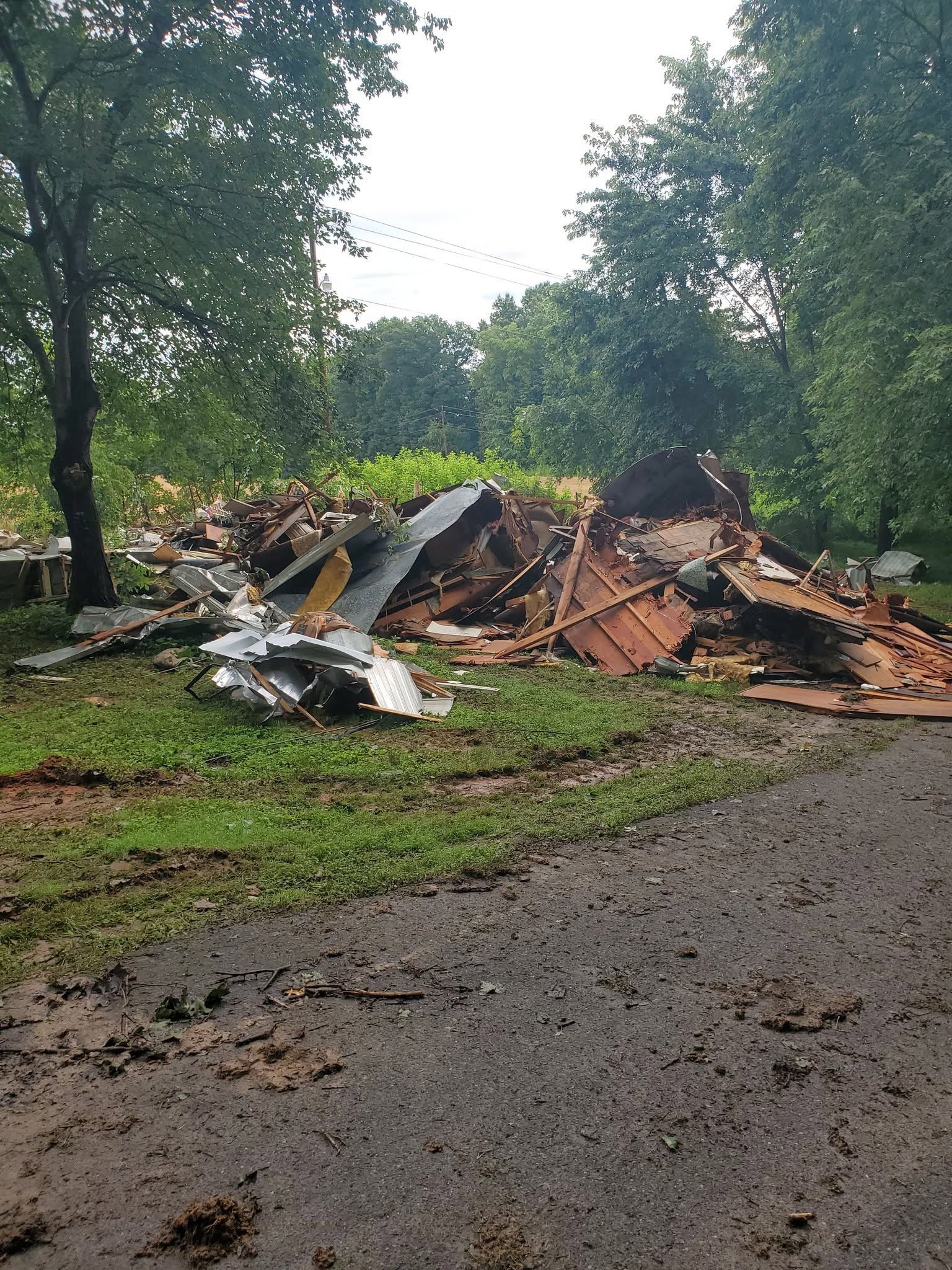 Collapsed house debris piled in a muddy, tree-lined yard after a storm or demolition.