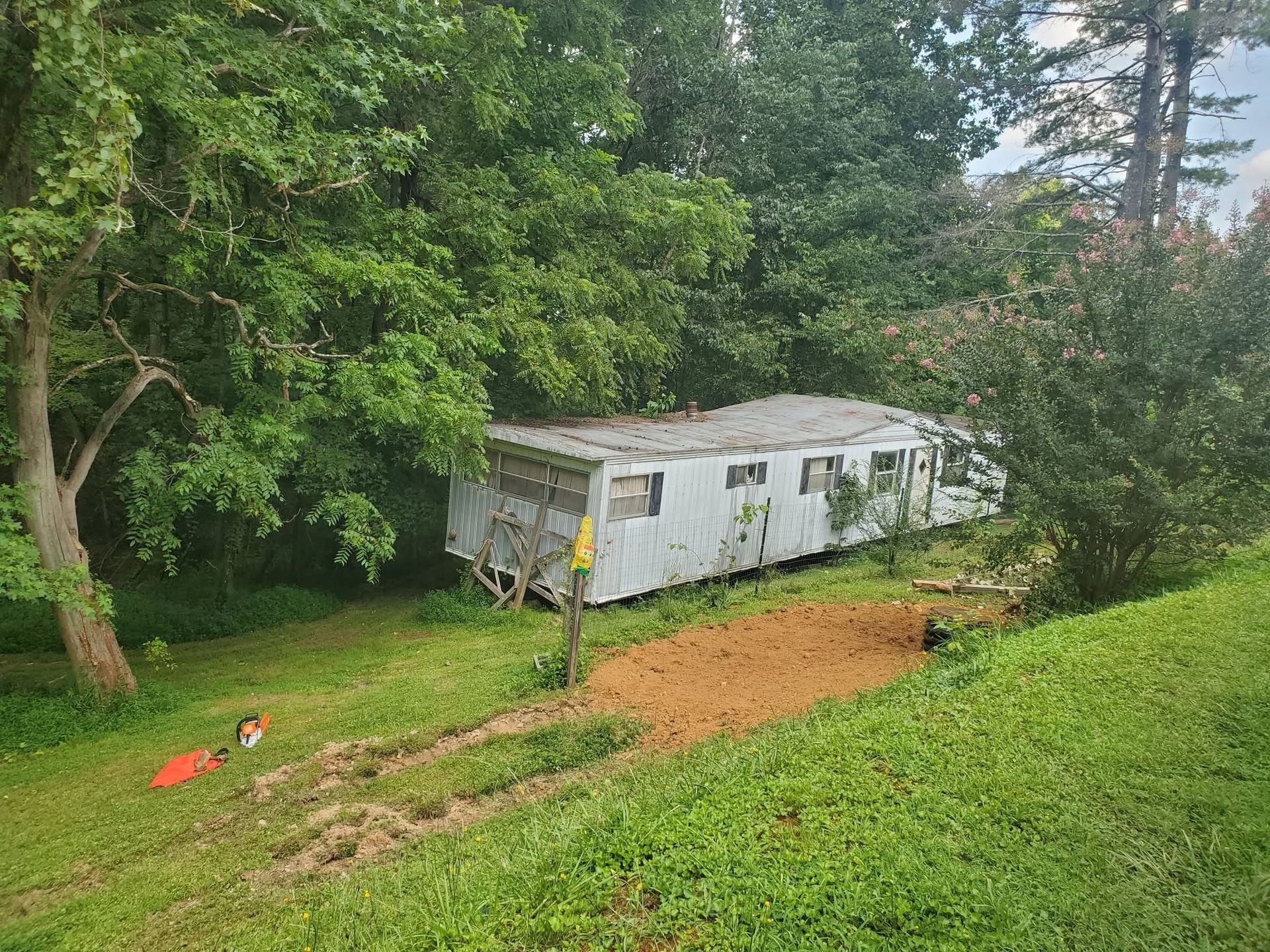 Old white trailer in a wooded yard, with a worker in yellow standing beside it on a dirt slope.