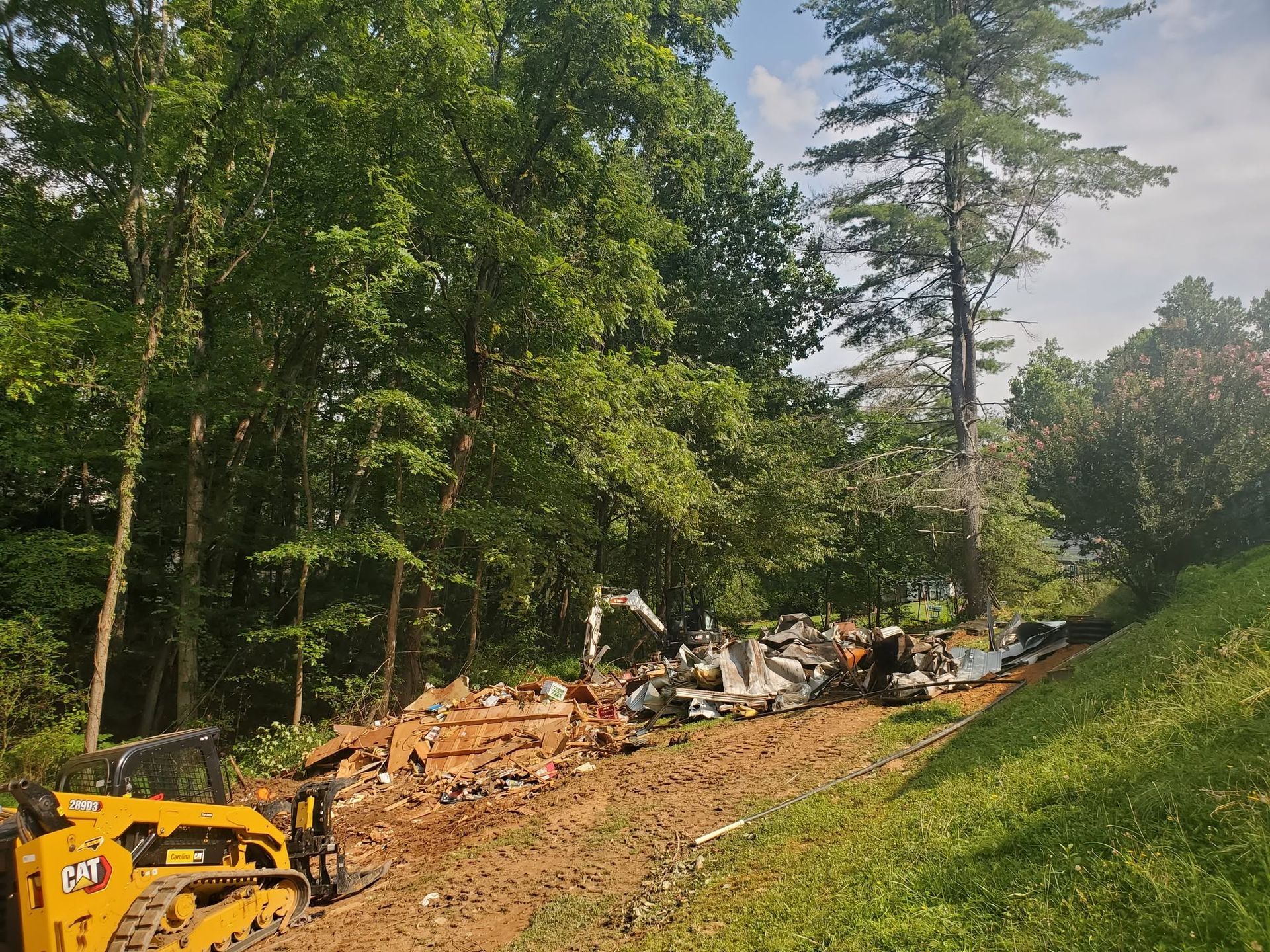Cleared wooded lot with excavator, fallen trees, and debris under green trees and a blue sky