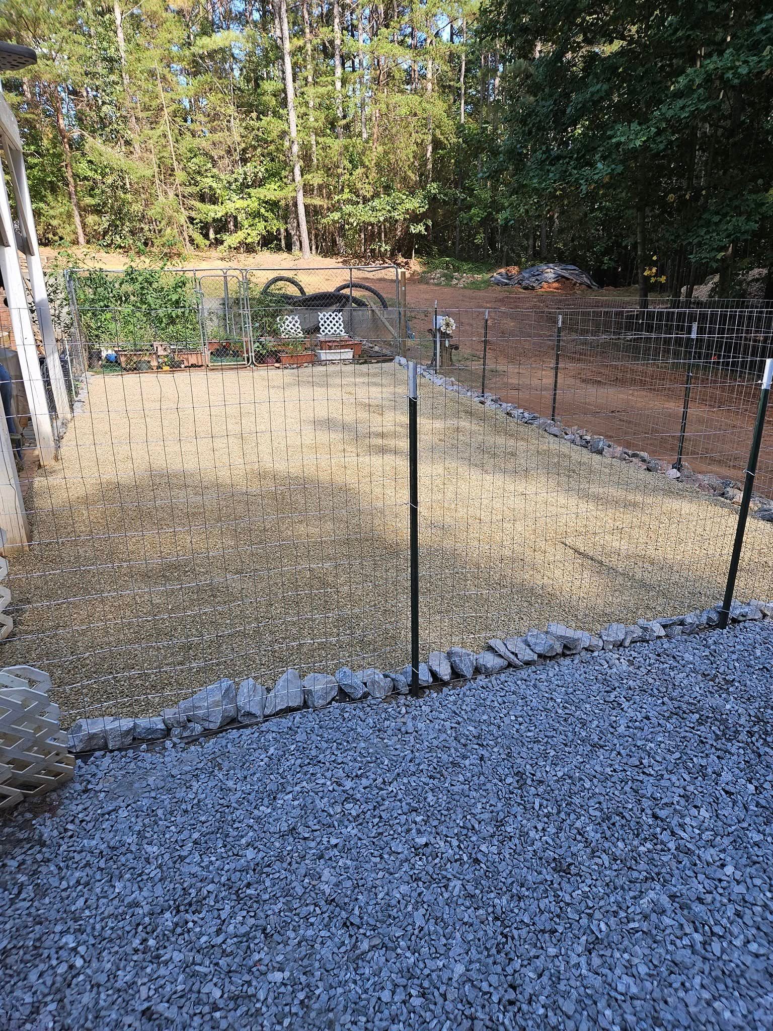 Backyard fenced in black posts, with gravel foreground and trees and shrubs in the background.