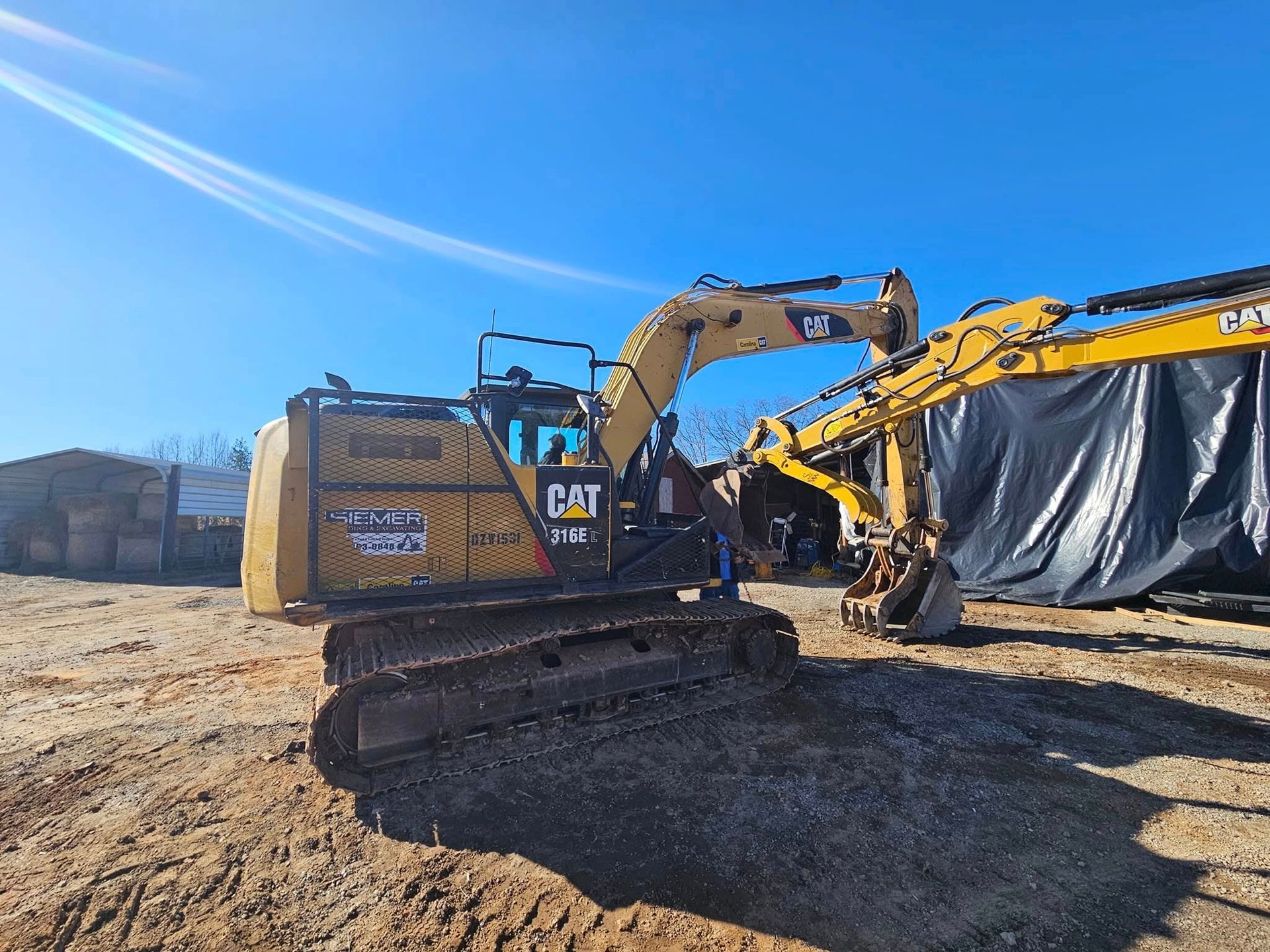 Yellow CAT excavator at a dirt worksite beside a tarped structure under a blue sky.