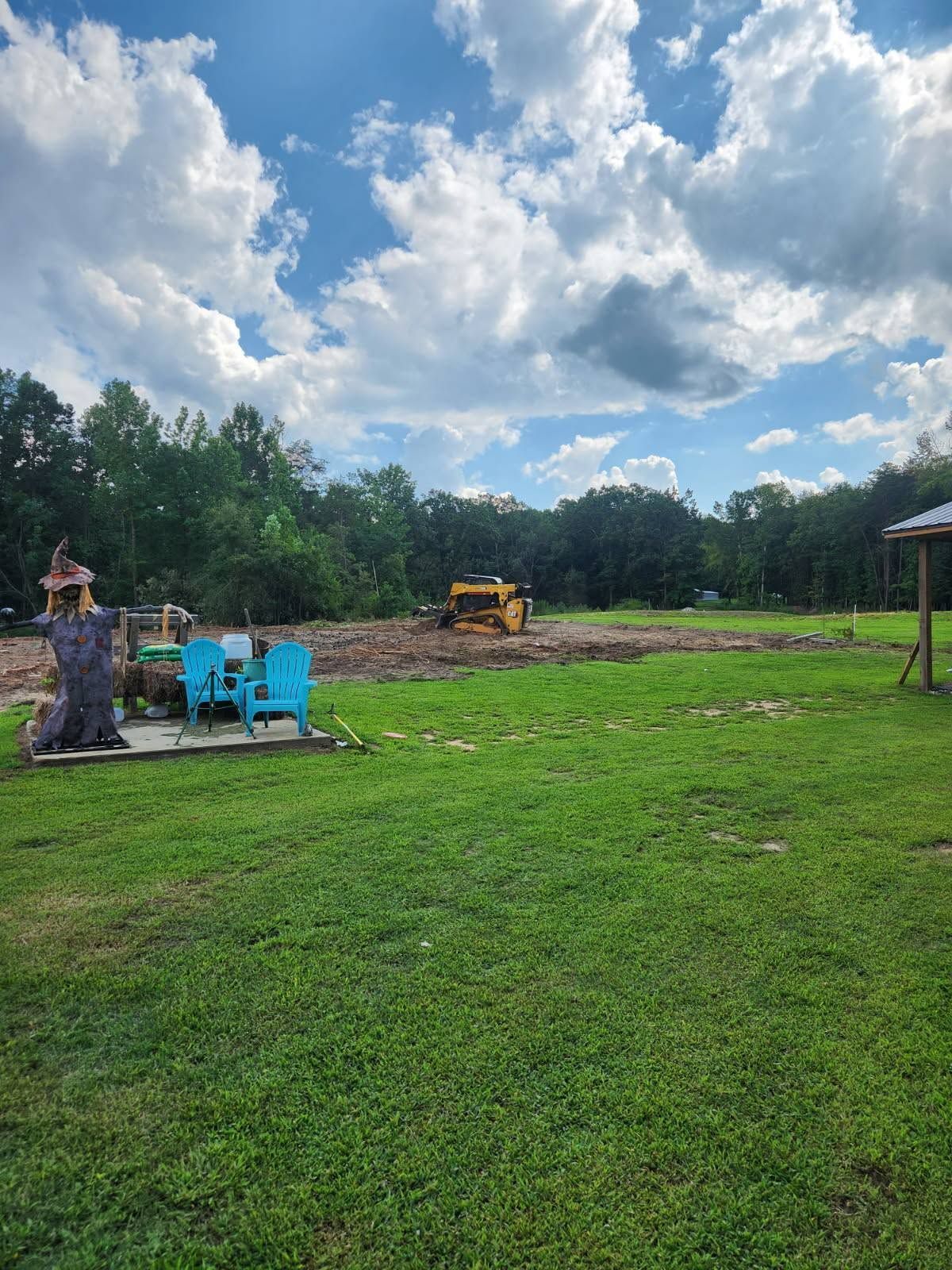 Grassy field with blue chairs, a tree stump, and a yellow bulldozer under a cloudy sky