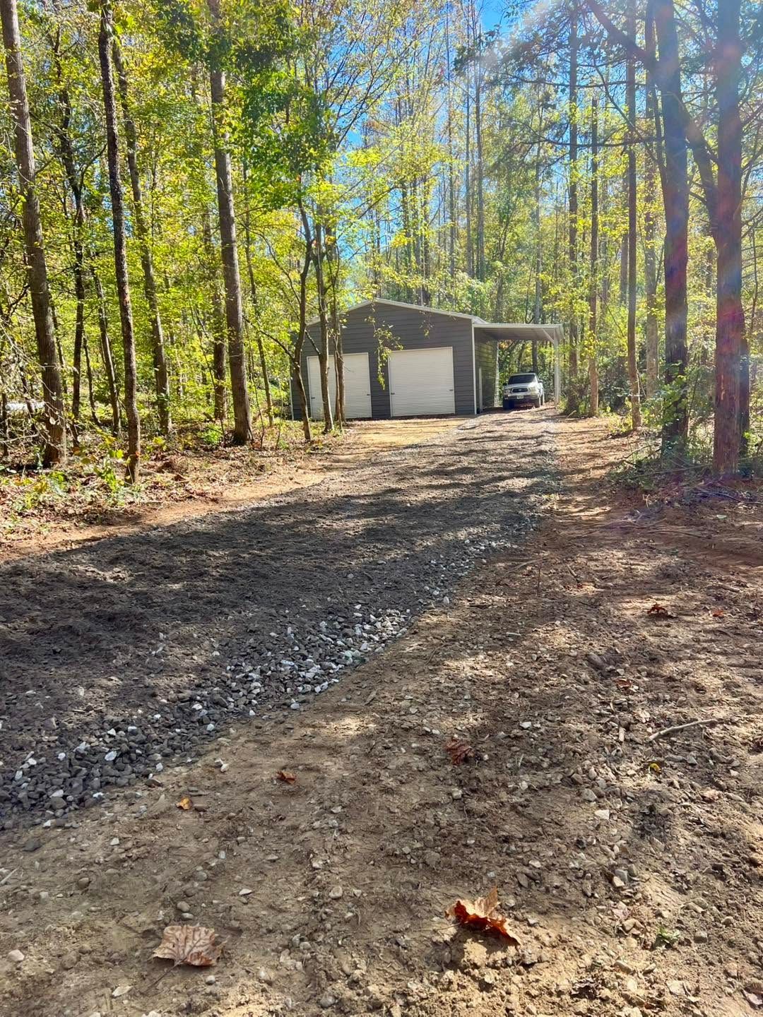 Gravel driveway leading to a small garage in a wooded area with a parked car and tall trees
