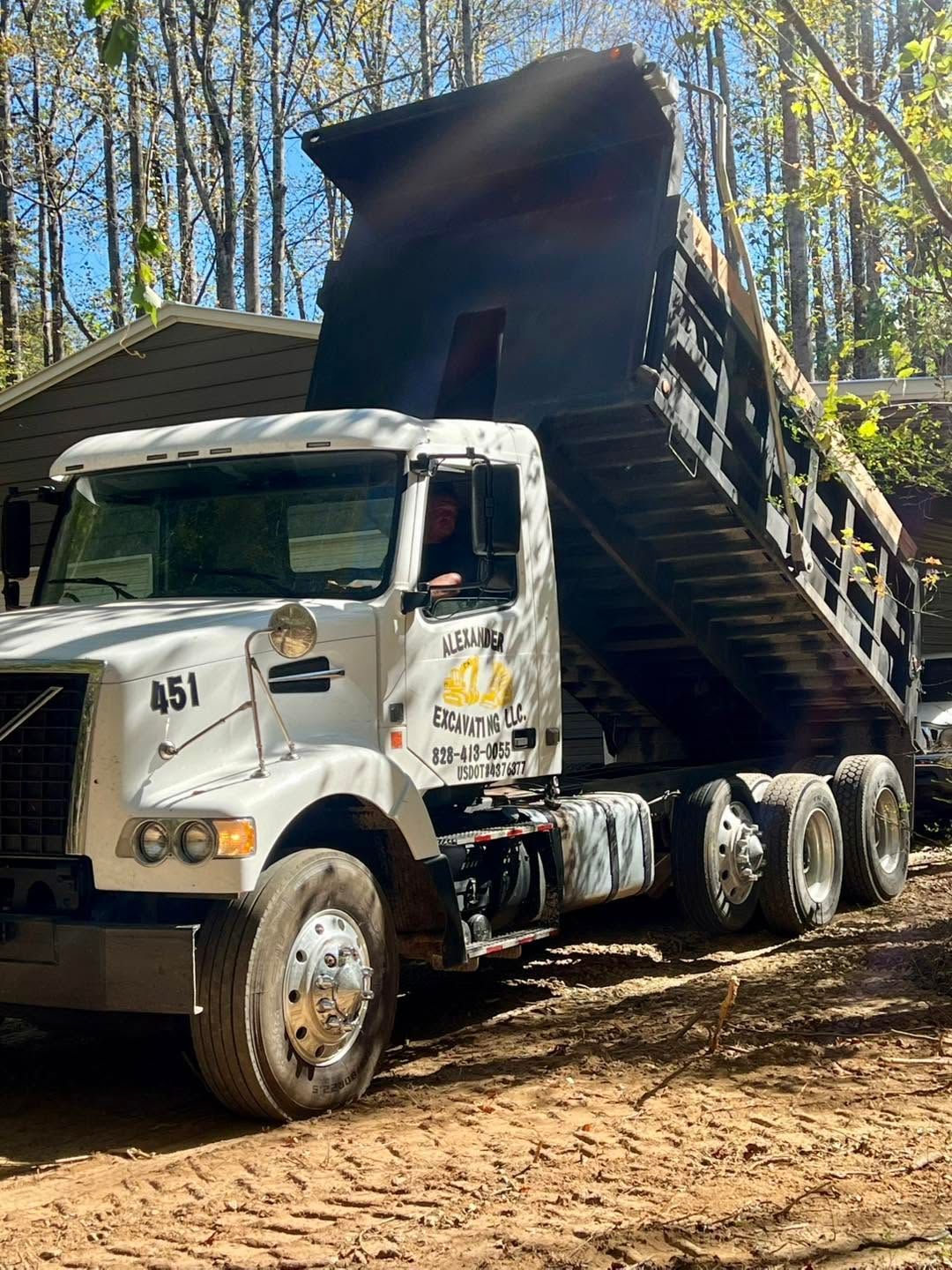 Dump truck unloading dark soil in a wooded area, with its bed raised steeply.