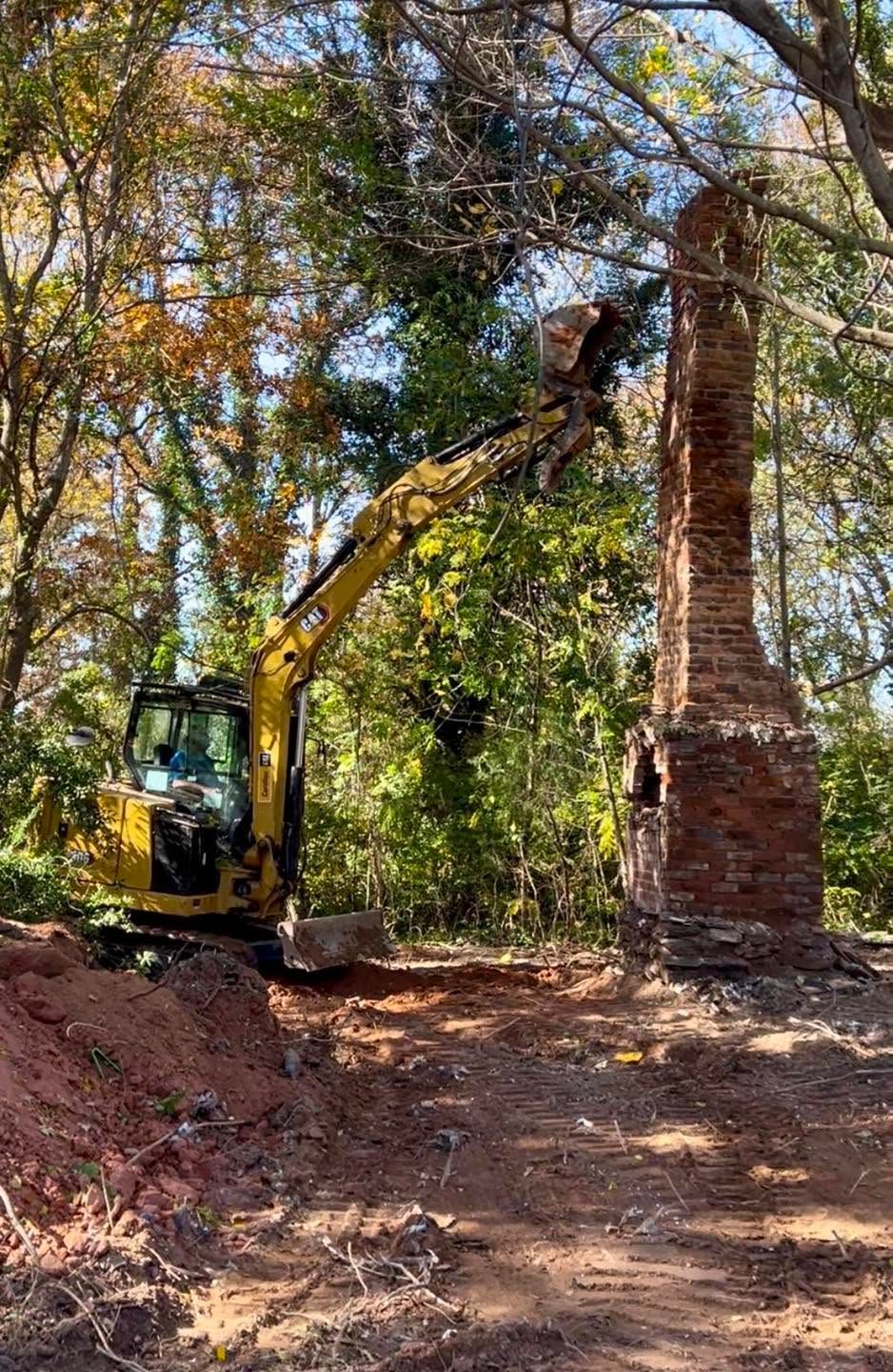 Excavator clearing a brick chimney in a wooded area, with trees and dirt surrounding the site