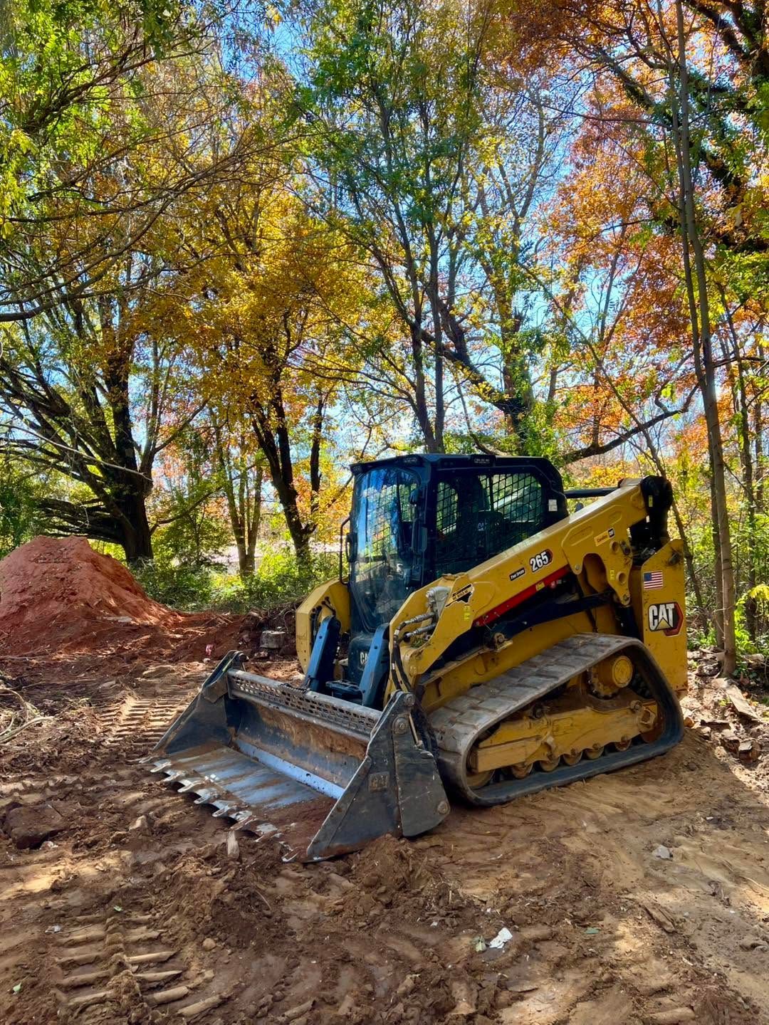Yellow skid-steer loader on a dirt site beside a mulch pile, with autumn trees in the background.
