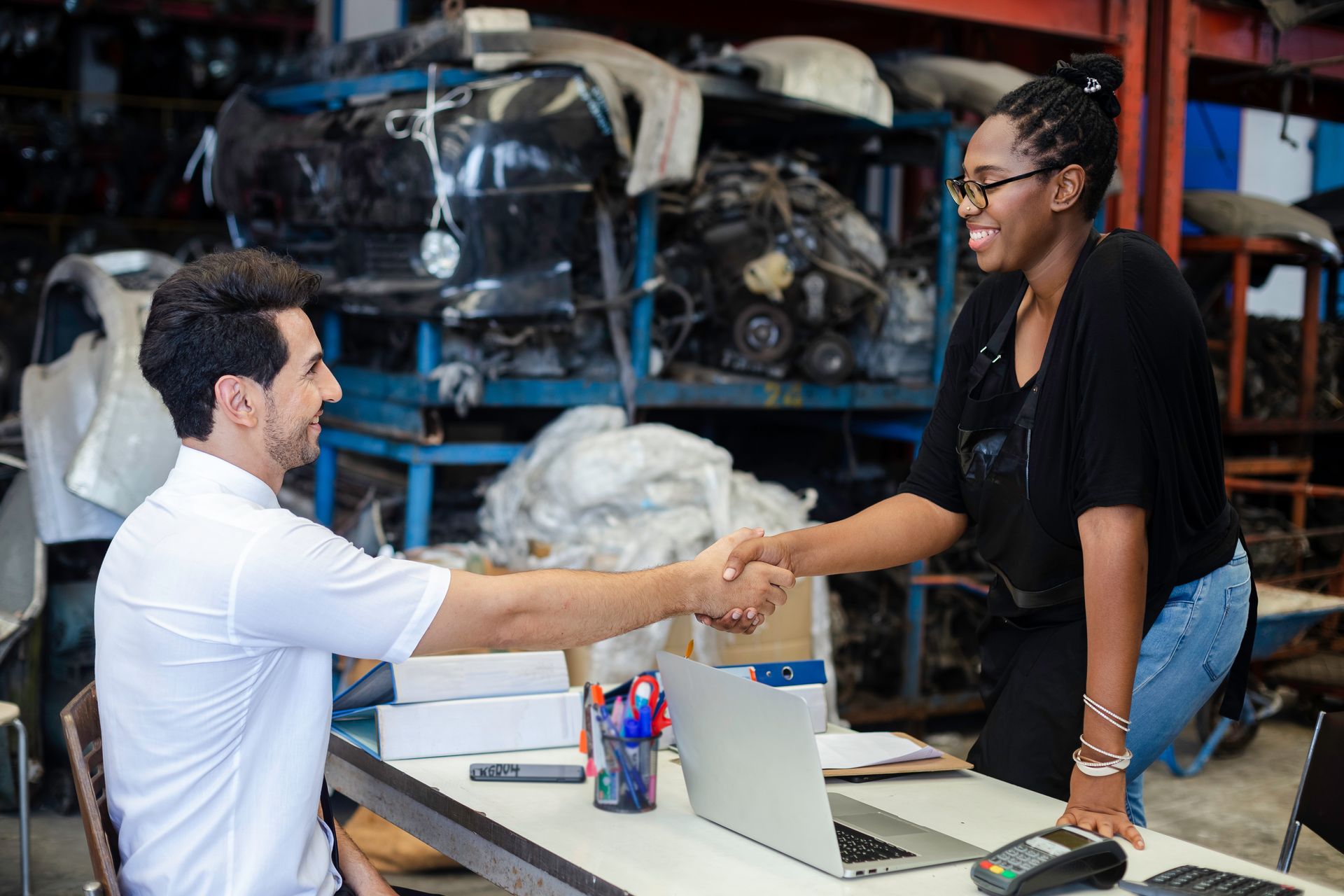 Man and woman shaking hands over a desk in a cluttered garage, smiling. Laptop and boxes present.