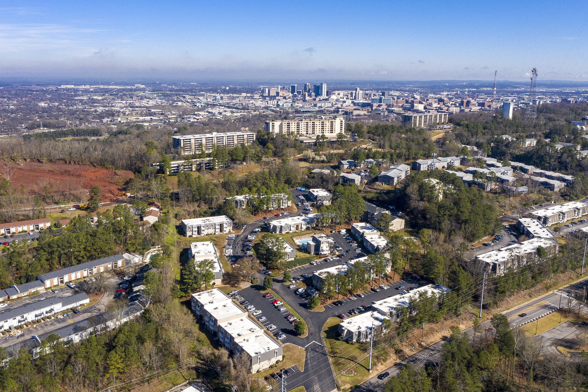 Aerial view of apartment complexes nestled in a tree-lined area, with a city skyline visible in the distant background.