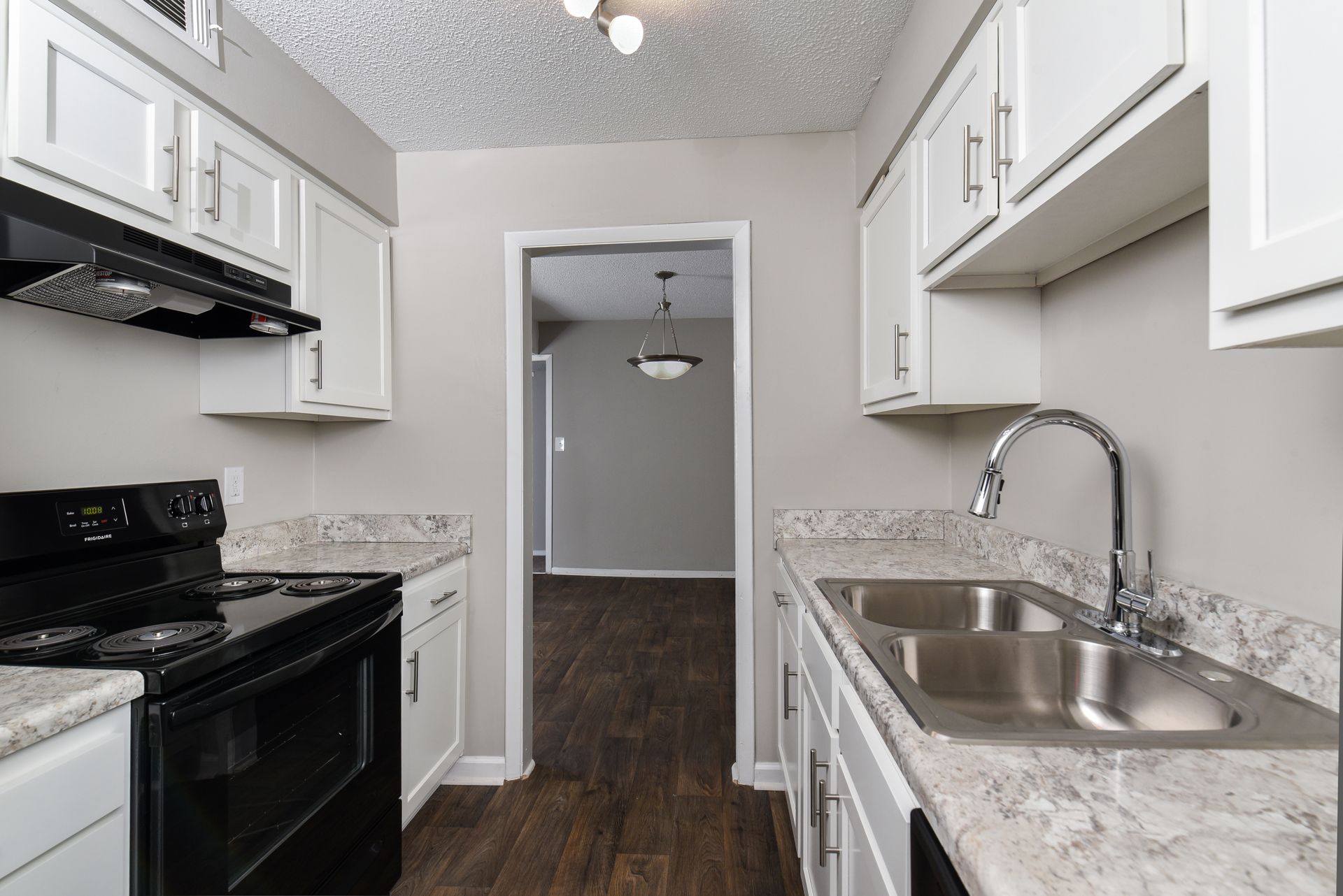 A galley kitchen with white cabinets, speckled stone countertops, a black stove, and a stainless steel double sink.