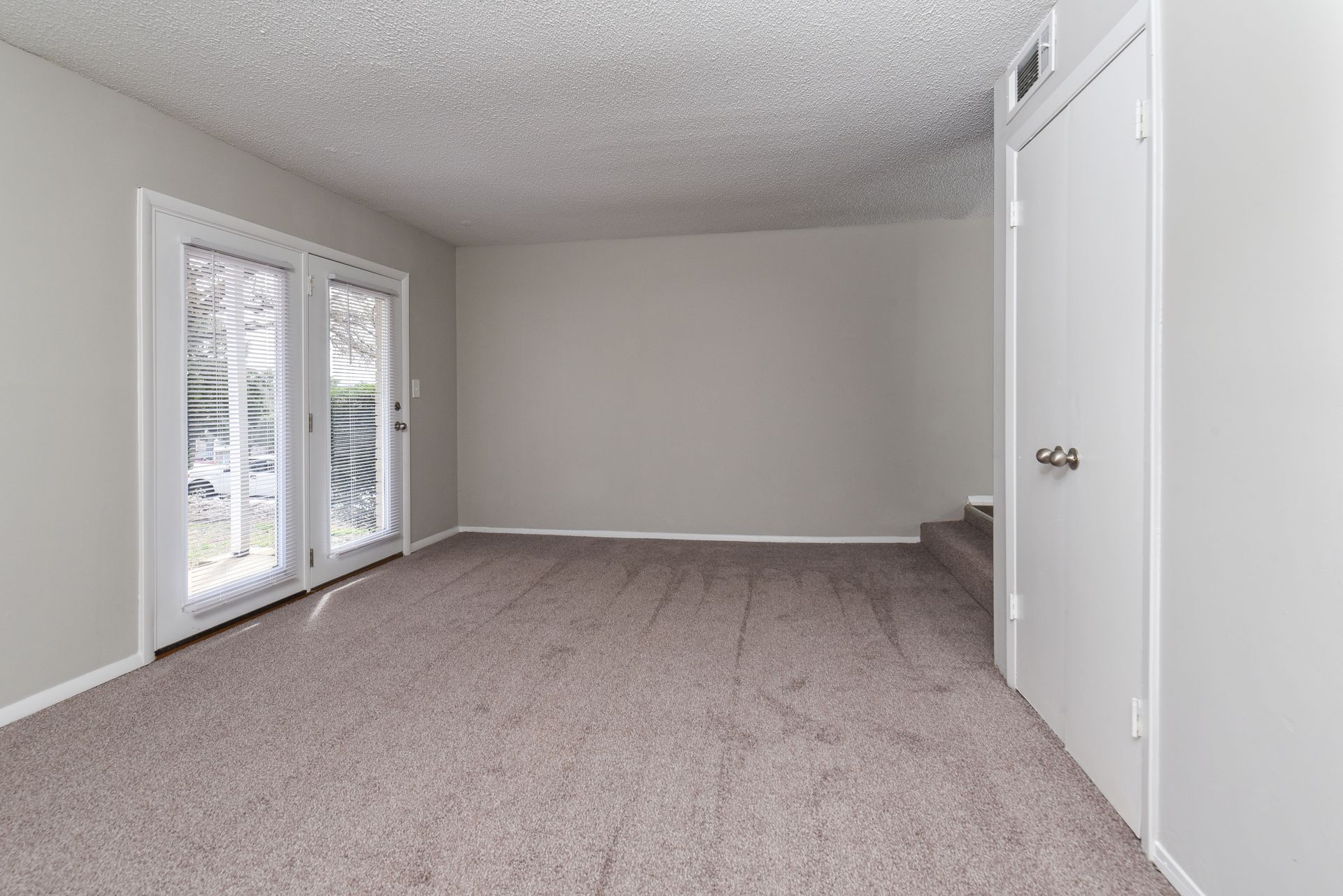 An empty room with gray carpet, plain walls, a set of glass double doors, and a closed white door on the right.