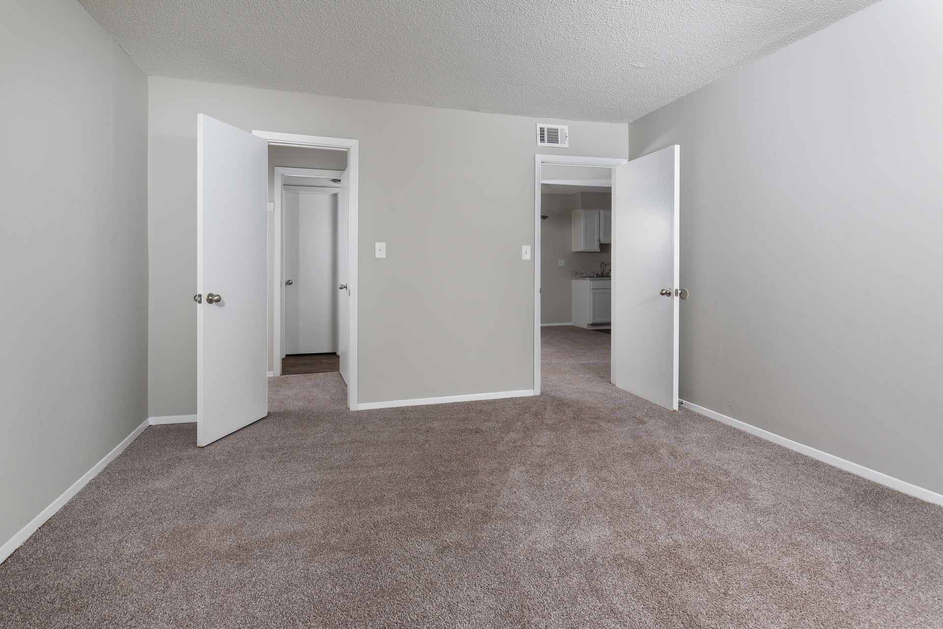 Empty room with gray walls, brown carpet, and two open white doors leading to an adjacent hallway and kitchen.
