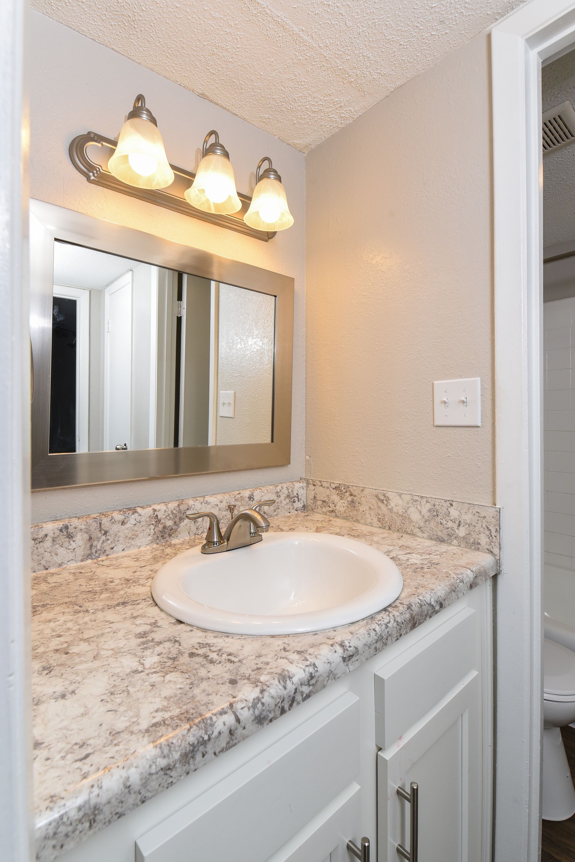 A bathroom vanity with a white sink, granite-style countertop, silver faucet, modern mirror, and a three-bulb light fixture.