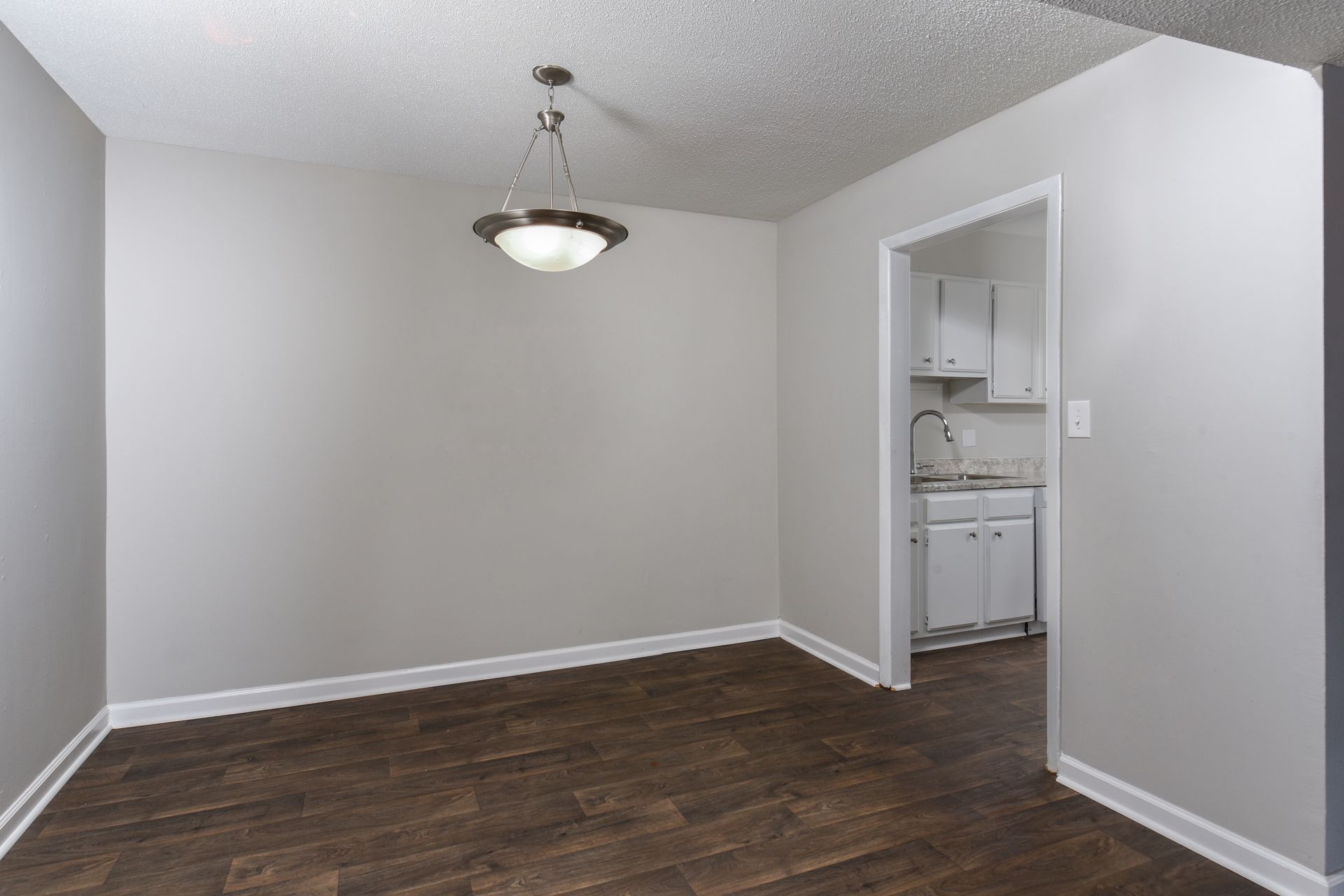 A small, empty dining room with grey walls, dark wood-look flooring, a pendant light, and a doorway leading to a kitchen.