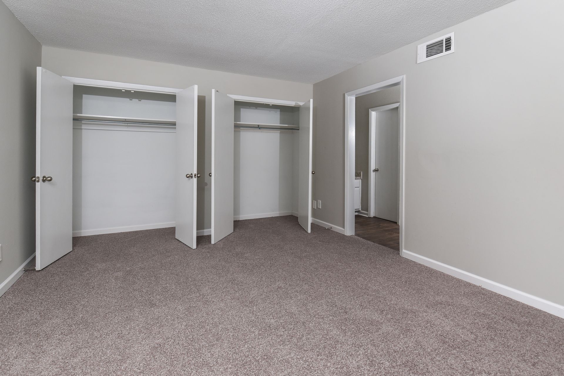 An empty bedroom with gray walls, beige carpet, popcorn ceiling, and two open white closets.