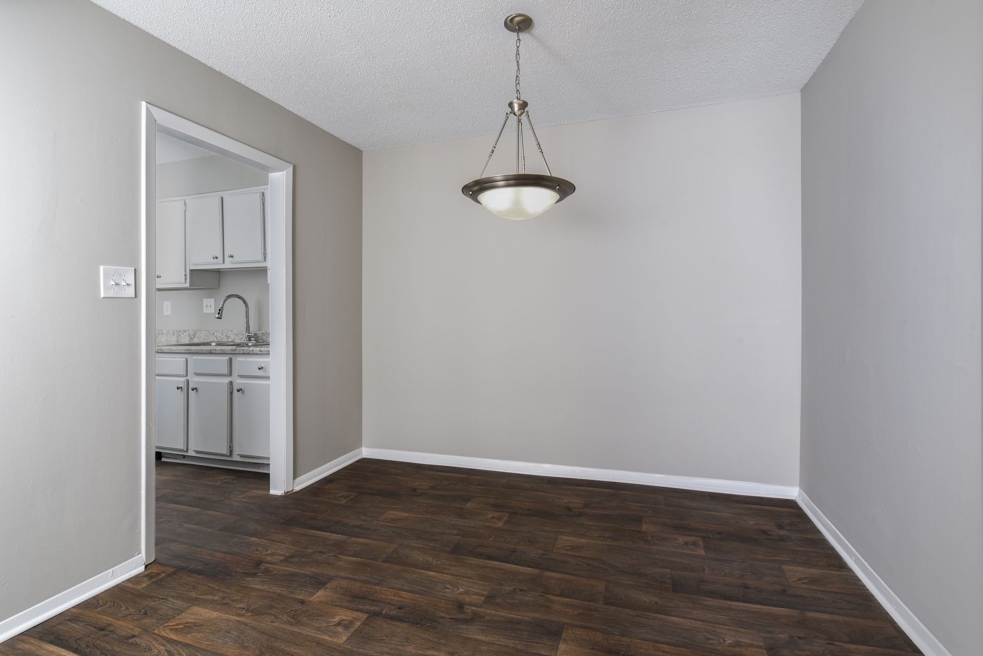 An empty dining room with dark wood flooring, light grey walls, and a hanging pendant light, leading to a kitchen.
