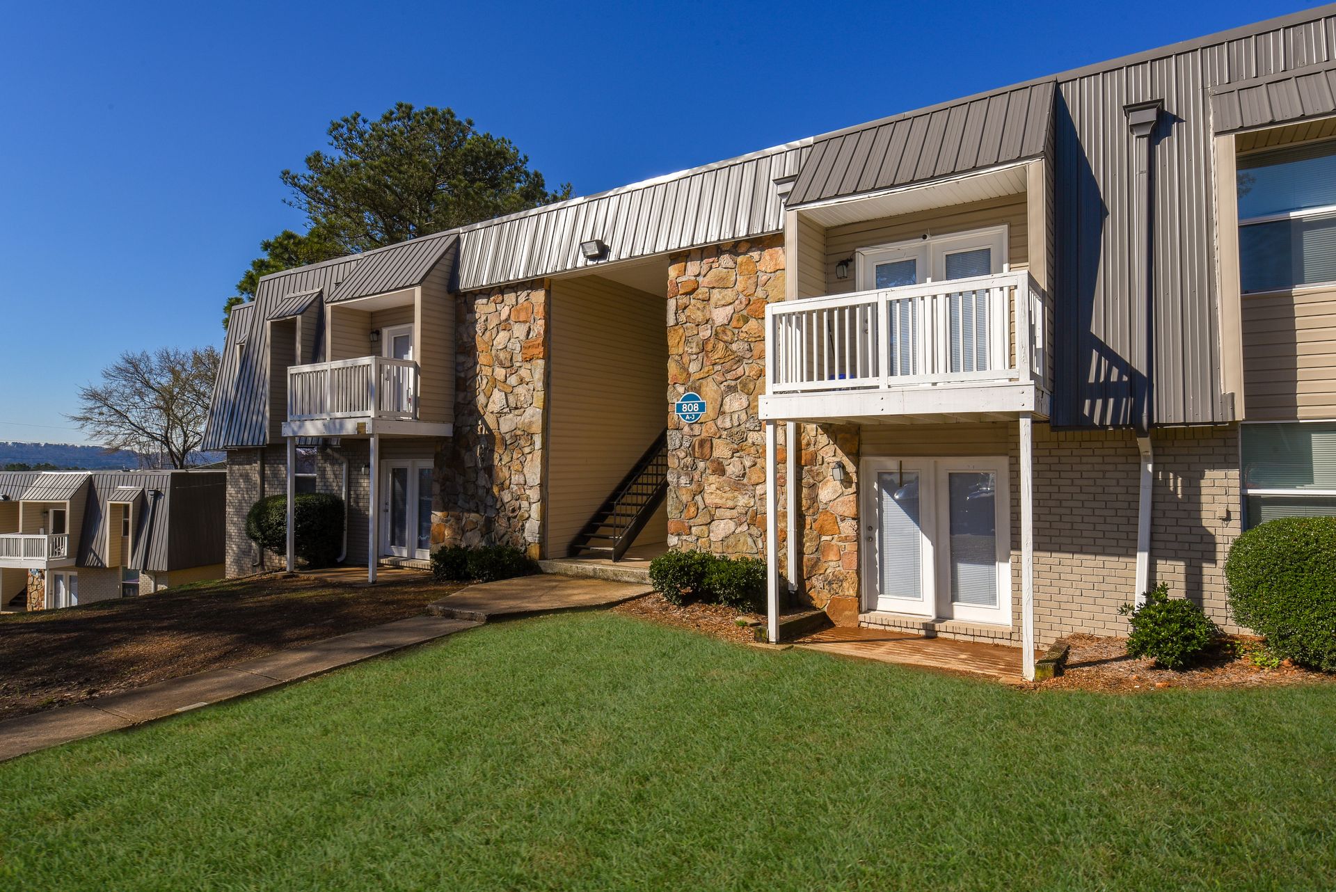 Two-story apartment building with stone masonry, white balconies, and green lawn under a clear blue sky.
