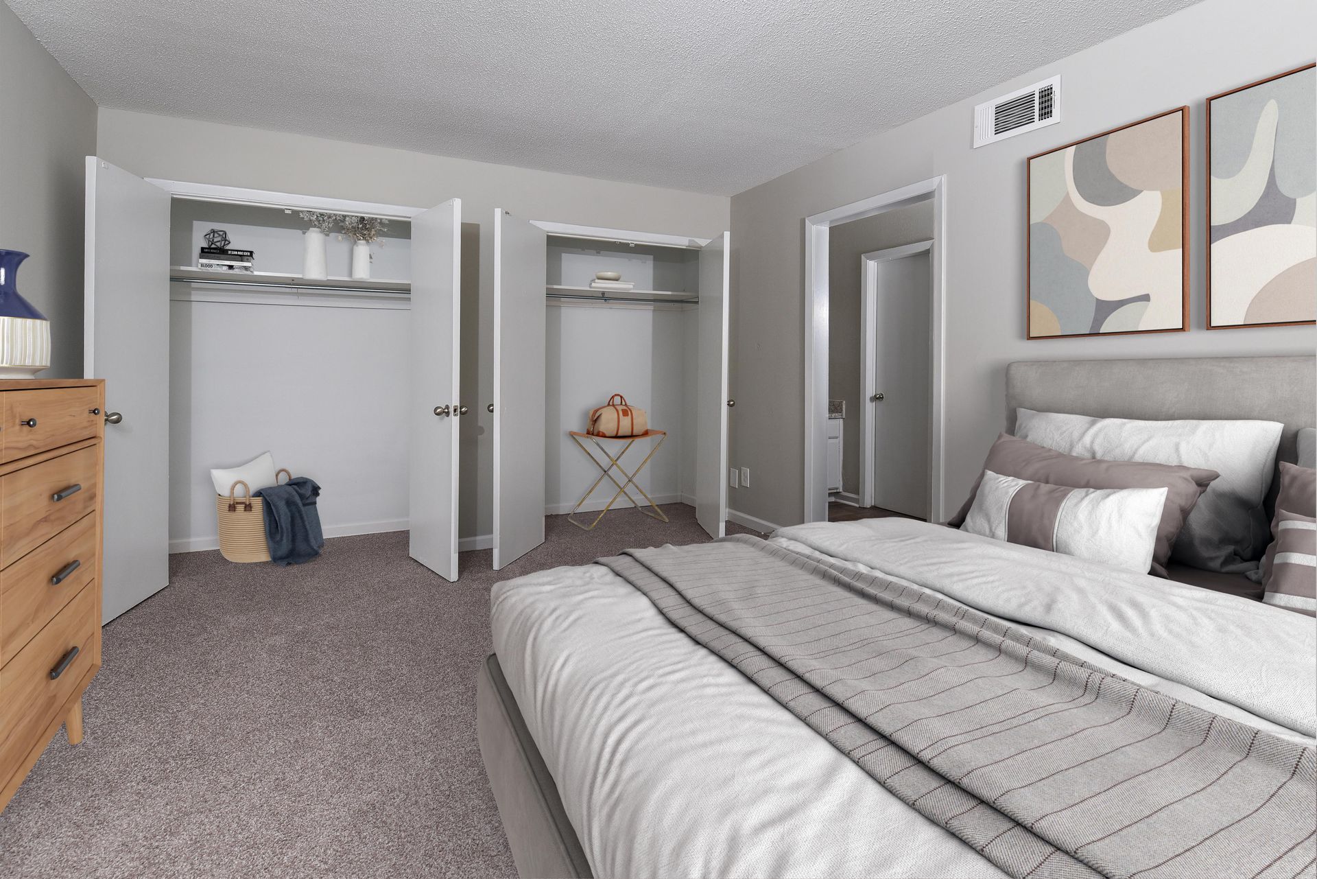 A bedroom with gray walls, carpet, a bed with a gray spread, wooden dresser, and two open closets with white doors.
