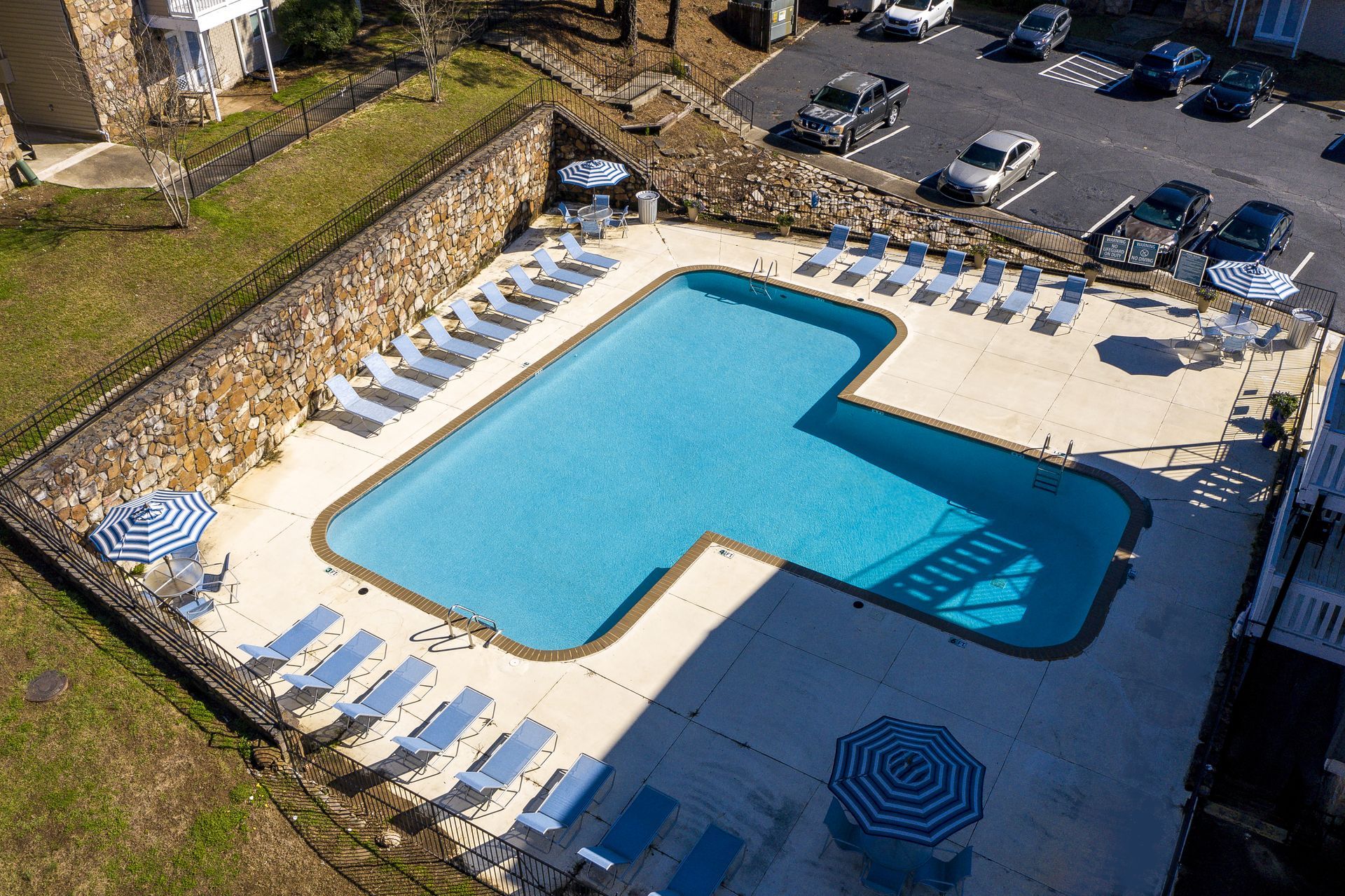 An aerial view of a T-shaped swimming pool surrounded by a patio with lounge chairs and umbrellas, near a parking lot.