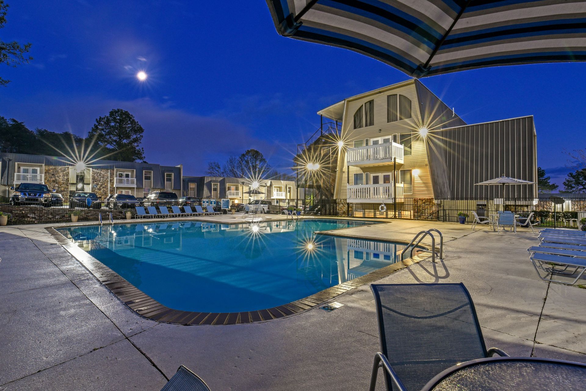 A multi-story hotel with a lit pool at night, under a dark blue sky with a visible moon.