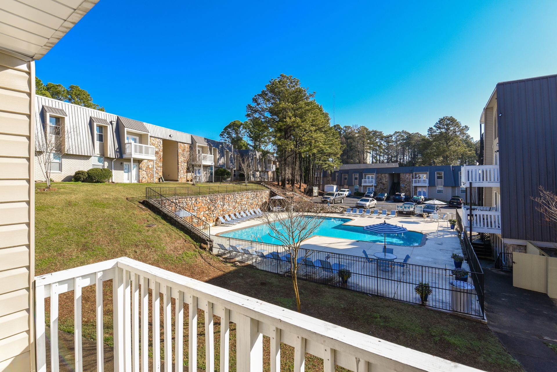 A sunny view from a balcony overlooking a swimming pool surrounded by a fence and residential apartment buildings.