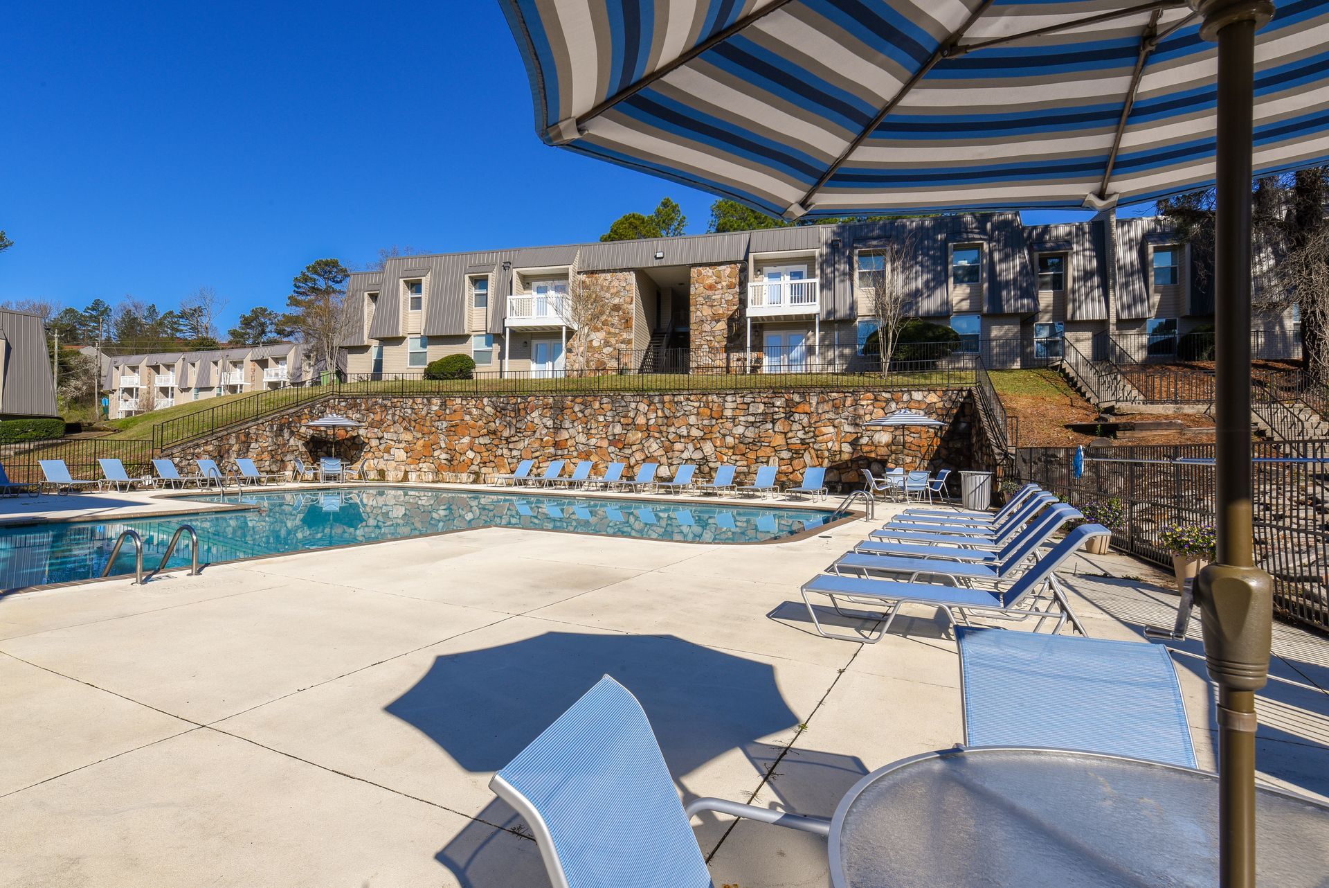 A swimming pool surrounded by lounge chairs and blue-striped umbrellas in front of a multi-story apartment complex.