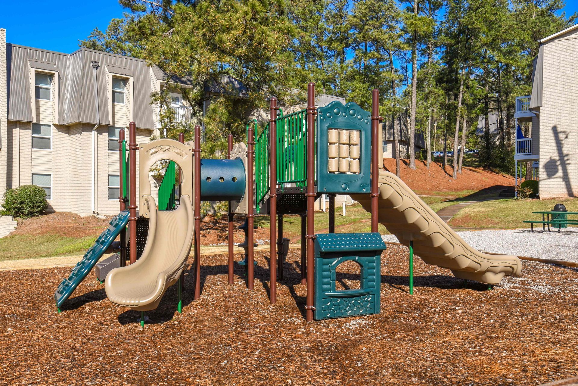 Playground structure with two tan slides, a blue tunnel, and green panels on wood mulch near an apartment building.