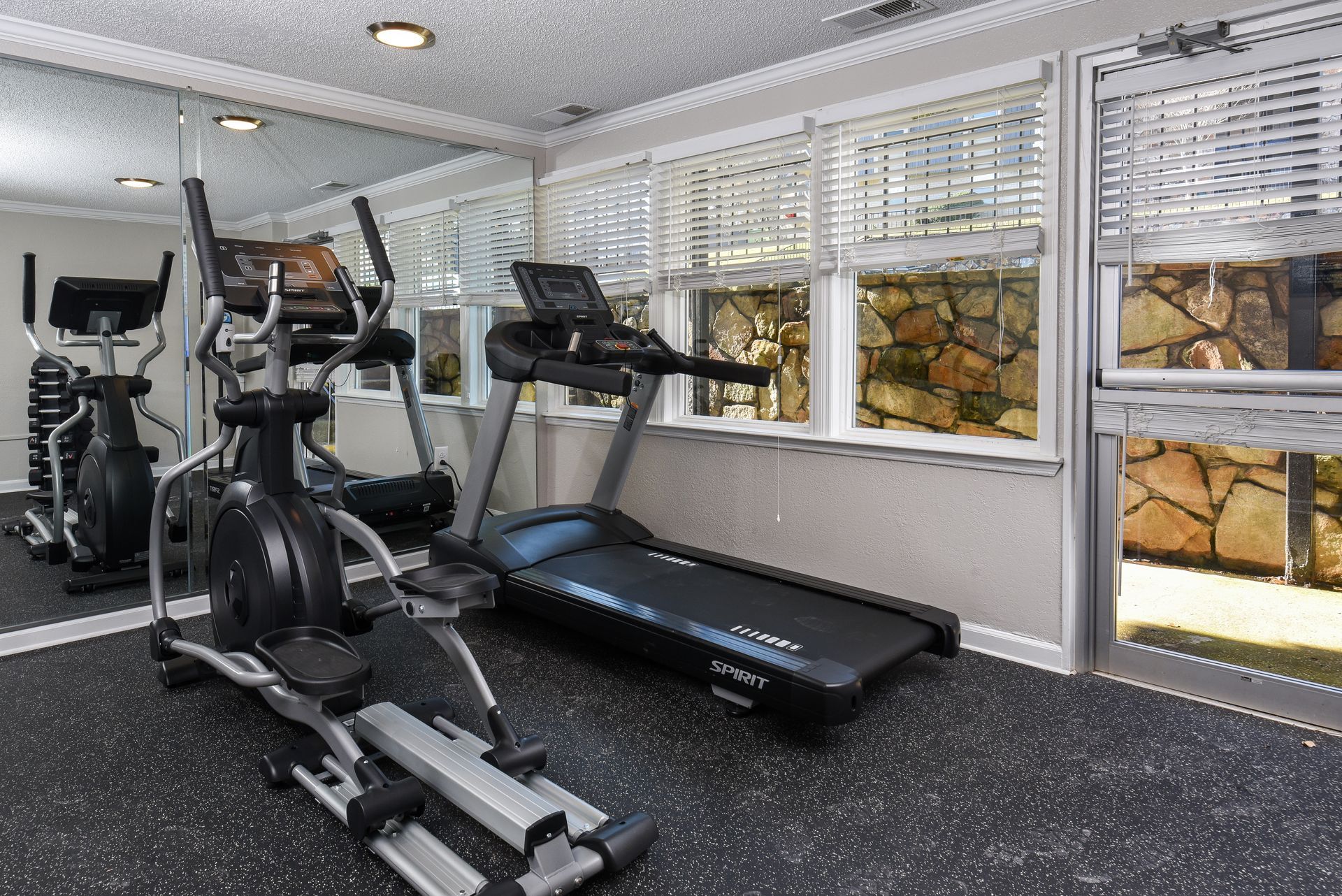A gym room featuring an elliptical machine and a treadmill, with a large mirror and windows looking onto a stone wall.