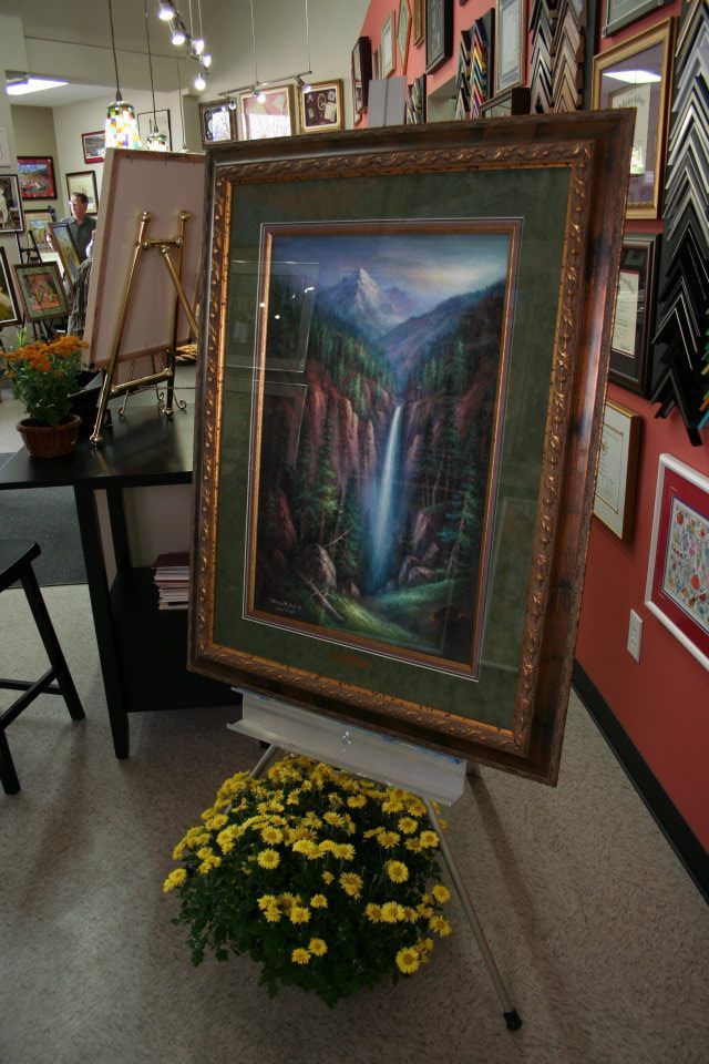 Framed landscape painting on an easel in an art gallery, with yellow flowers at the base